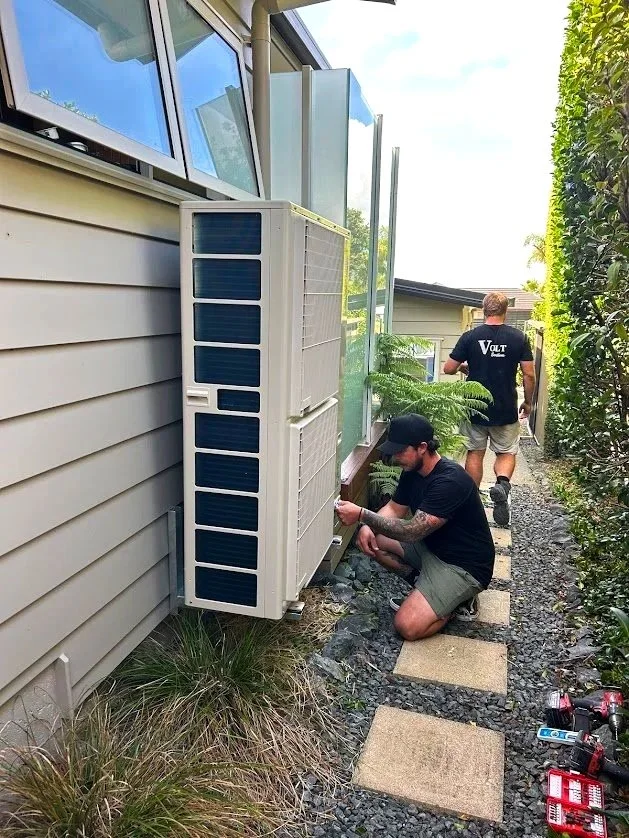 Volt Solutions electricians working on an air conditioning unit outside a house, one is kneeling and the other is walking away, with tools laying on the ground, next to a path of stepping stones.
