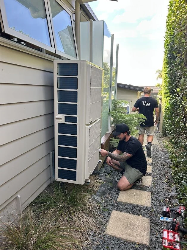 Two men working on outdoor air conditioning unit outside a house, one crouching and using tools, the other walking away.
