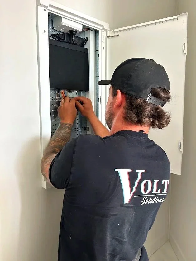 Volt Solutions electrician wearing a black cap and a black t-shirt labeled Volt Solutions working inside an open electrical cabinet, installing or repairing wiring.