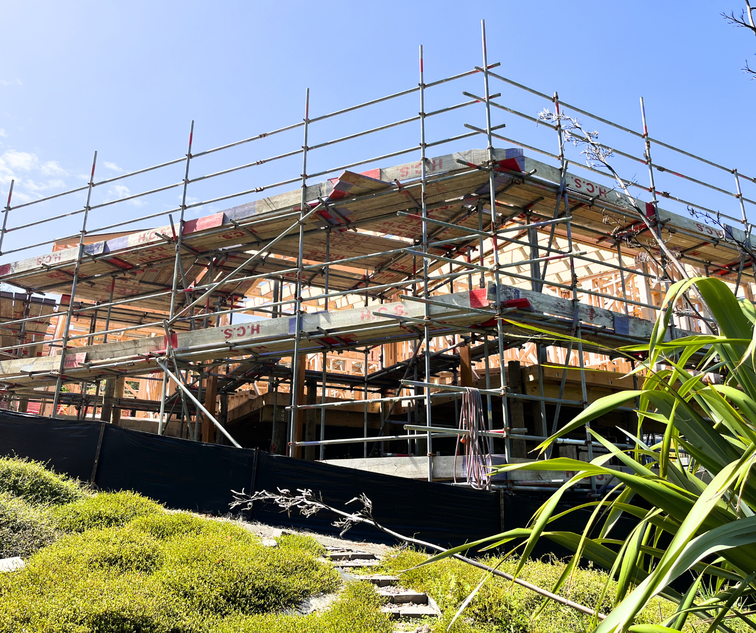 Construction site with a wooden framework and metal scaffolding under a clear blue sky, surrounded by green plants.