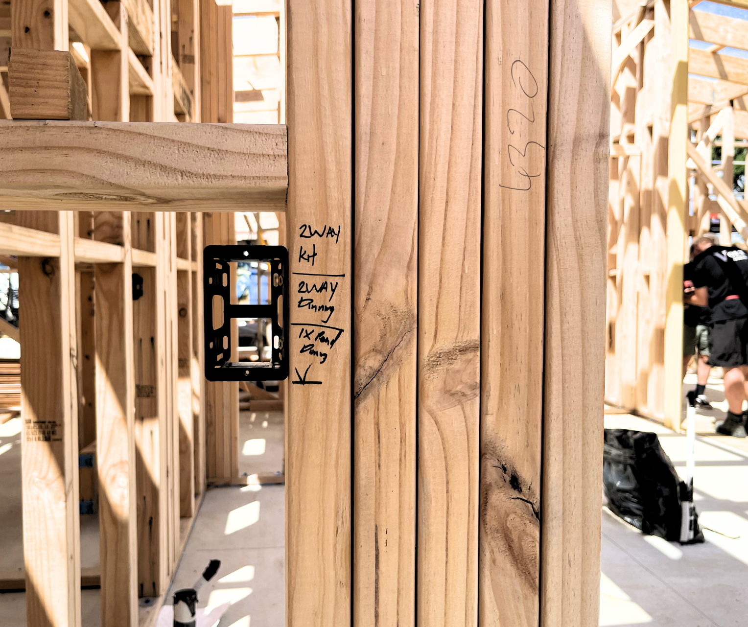 Interior view of a house under construction with wooden framing. Handwritten notes and measurements are displayed on the wood studs. In the background, a worker is visible working on the structure.  Volt Solutions electrician installed the electrics.