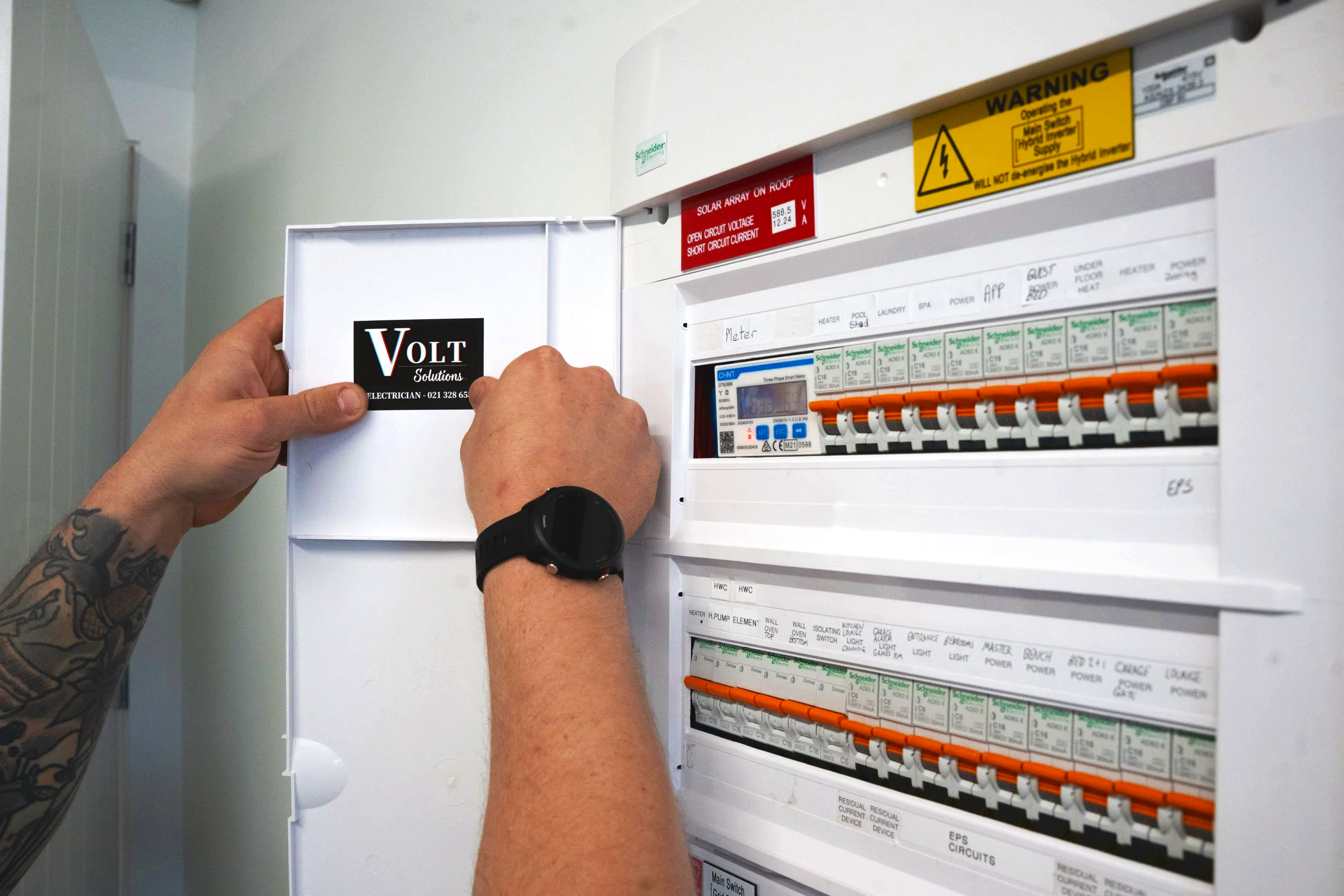Someone opening an electrical panel labeled Volt Solutions, with a tattooed arm and a black watch, in an indoor setting.