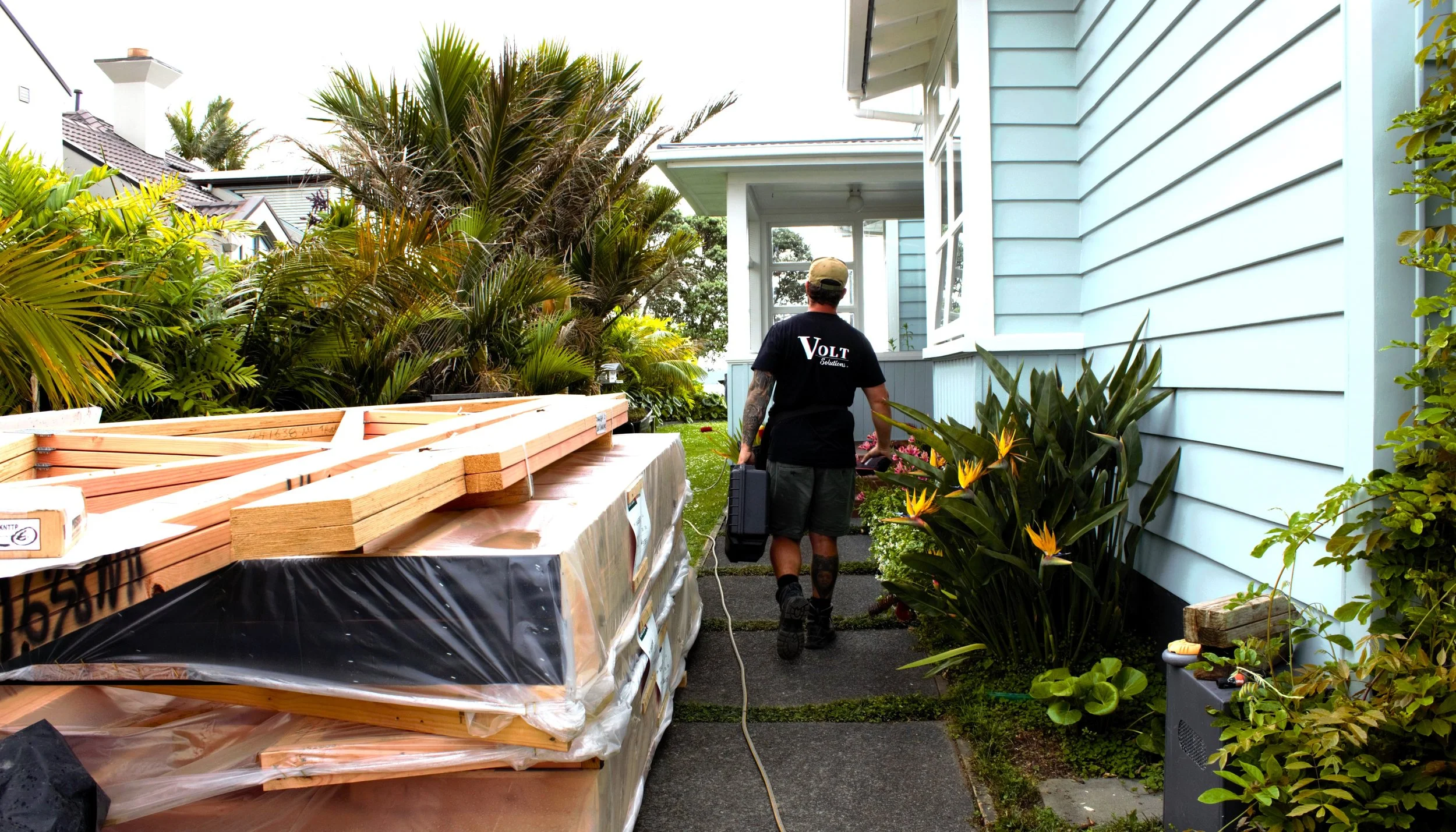A Volt Solutions electrician .walking along a pathway outside a house, carrying a toolbox, with stacked lumber on the left and lush tropical plants on the right.