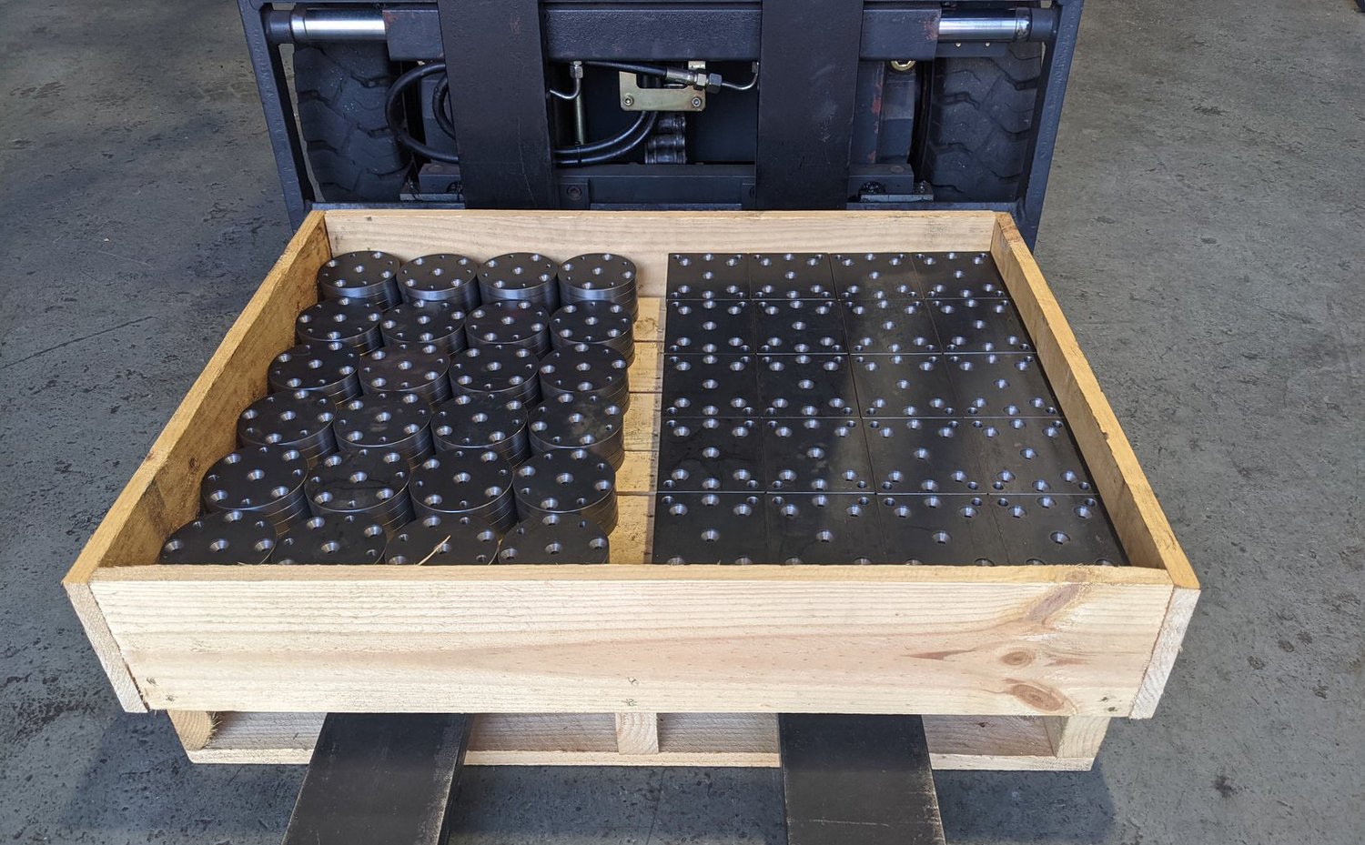 Wooden crate on a forklift holding metal parts with drilled holes, including circular and rectangular shapes.