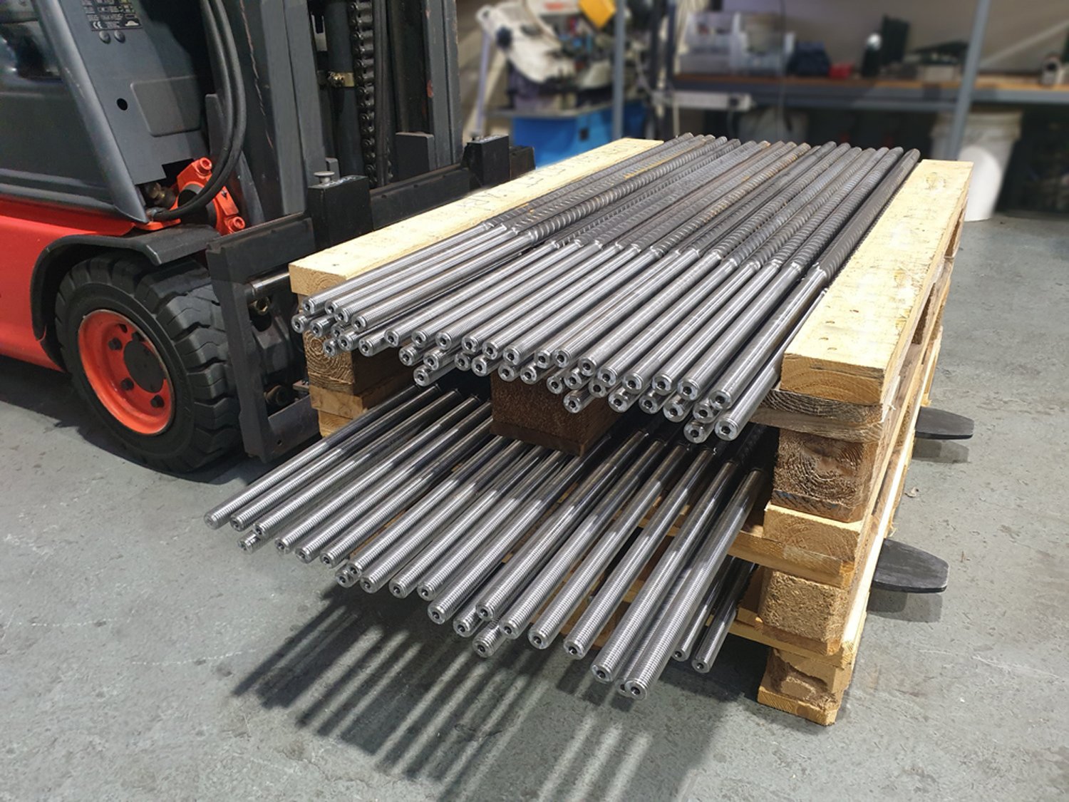Stacked metal rods on a wooden pallet with a forklift in a warehouse setting.