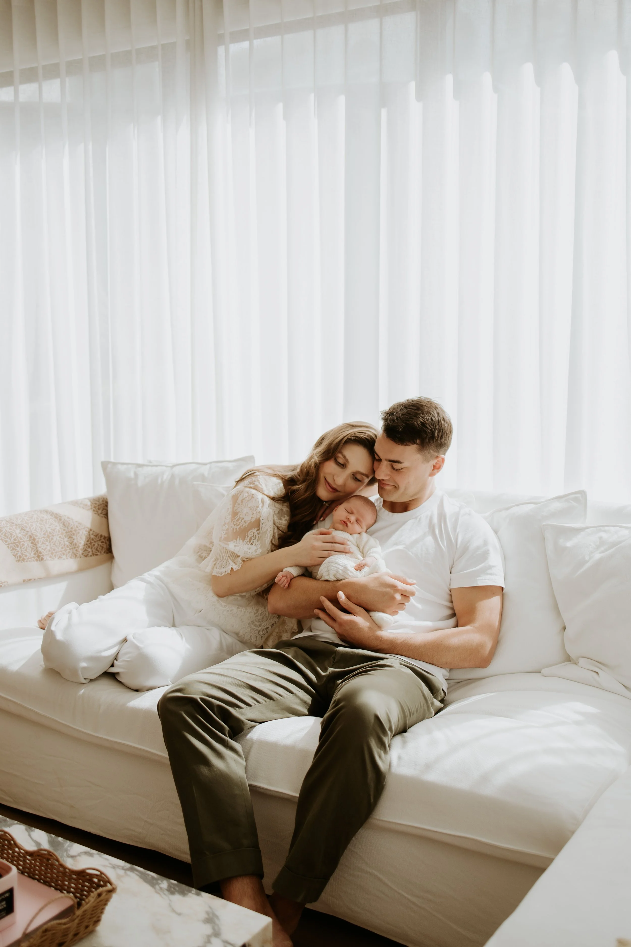 A young family sitting on a white couch, holding a newborn baby, in a bright room with white curtains.