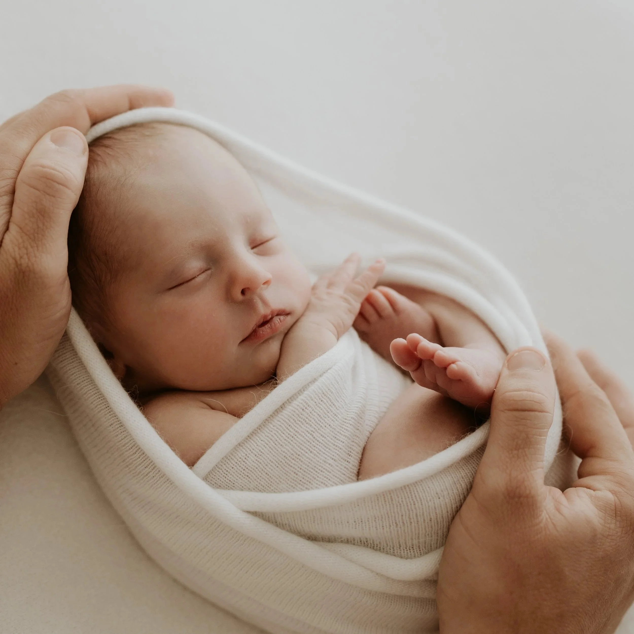Close-up of a sleeping newborn baby wrapped in a white cloth, gently cradled in adult hands.