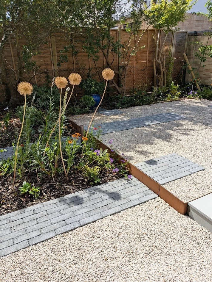 A backyard garden with flowering plants and a paved pathway, enclosed by a wooden fence and surrounded by trees and shrubs.