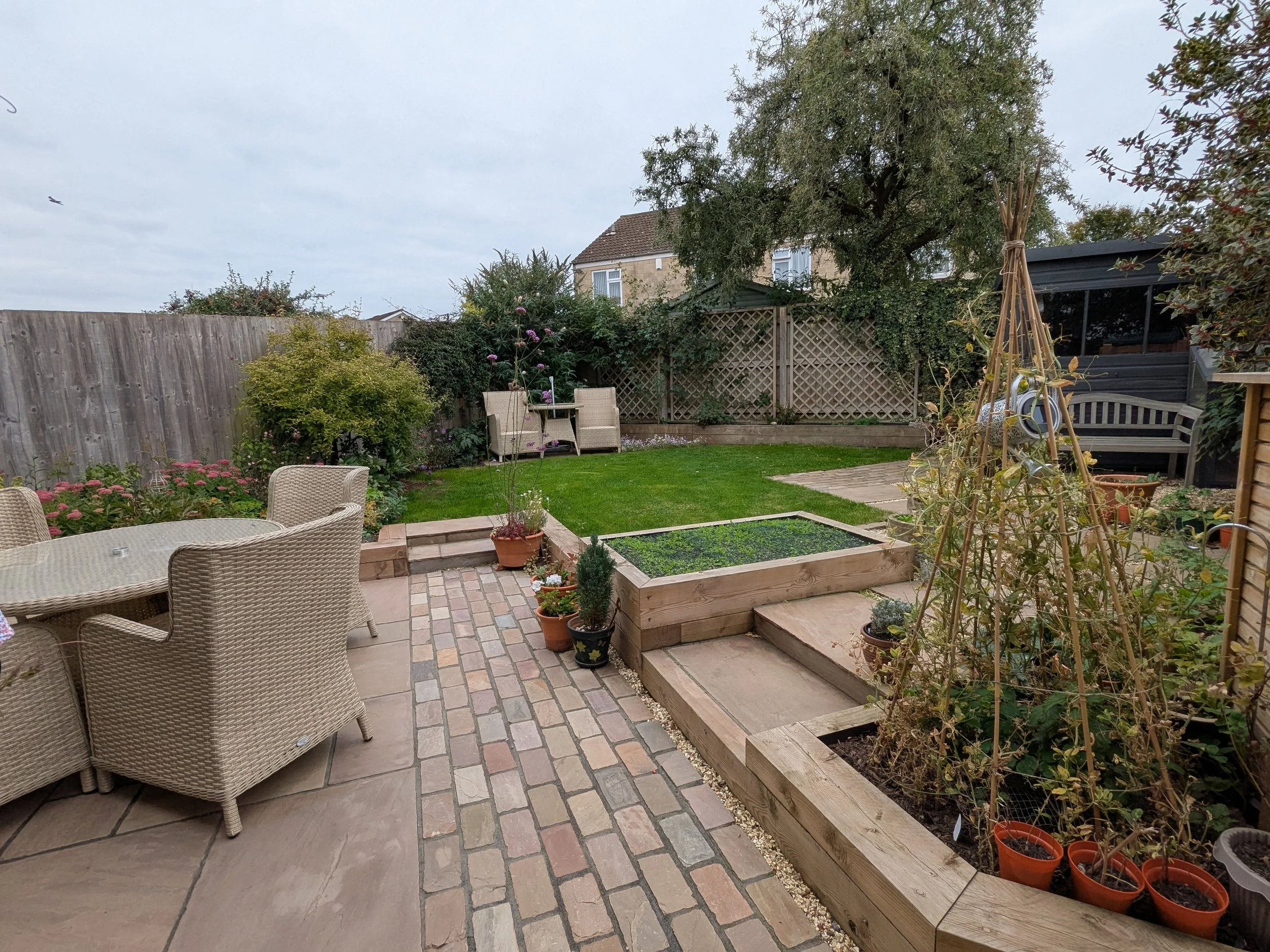 Backyard garden with patio, wicker chairs and table, potted plants, a small grassy lawn, trees, and a wooden fence.