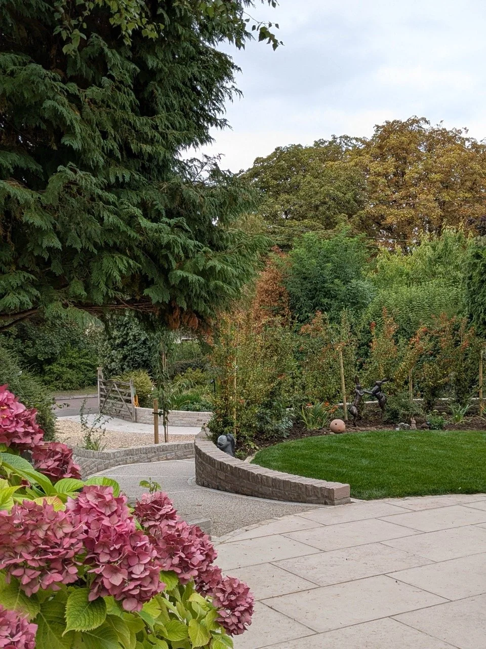 A well-maintained garden with a paved patio, lush green grass, colorful flowers, and various trees and shrubs. Decorative sculptures are visible among the plants.