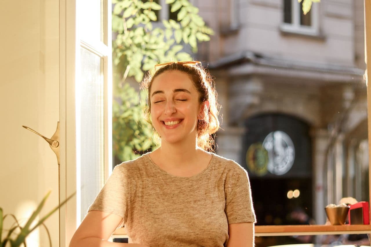 a-happy-woman-in-gray-shirt-smiling-while-sitting-near-glass-window