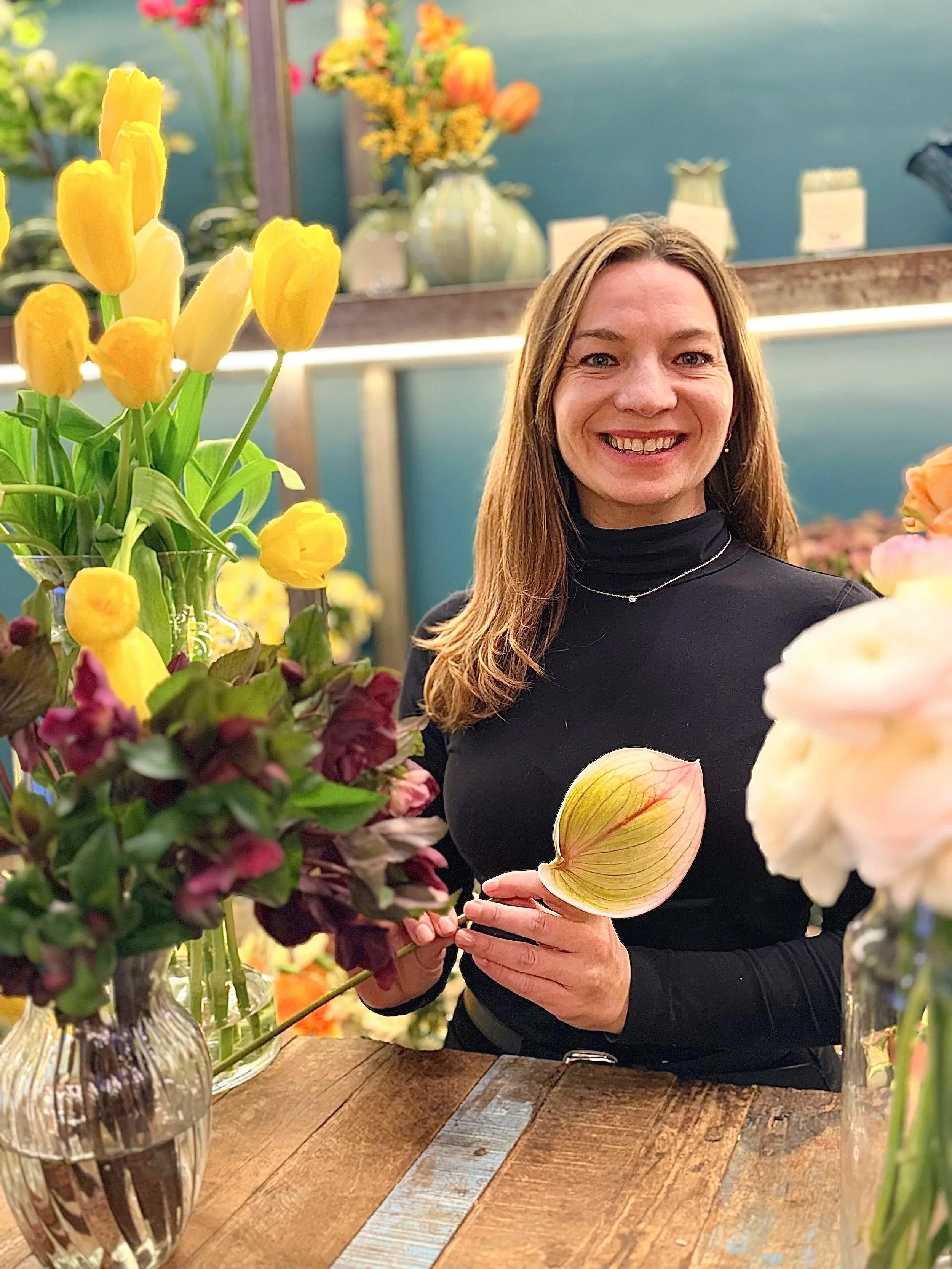 Mujer sonriente en una tienda de flores, rodeada de arreglos florales con tulipanes amarillos y otras flores de colores, sosteniendo una flor grande en la mano.