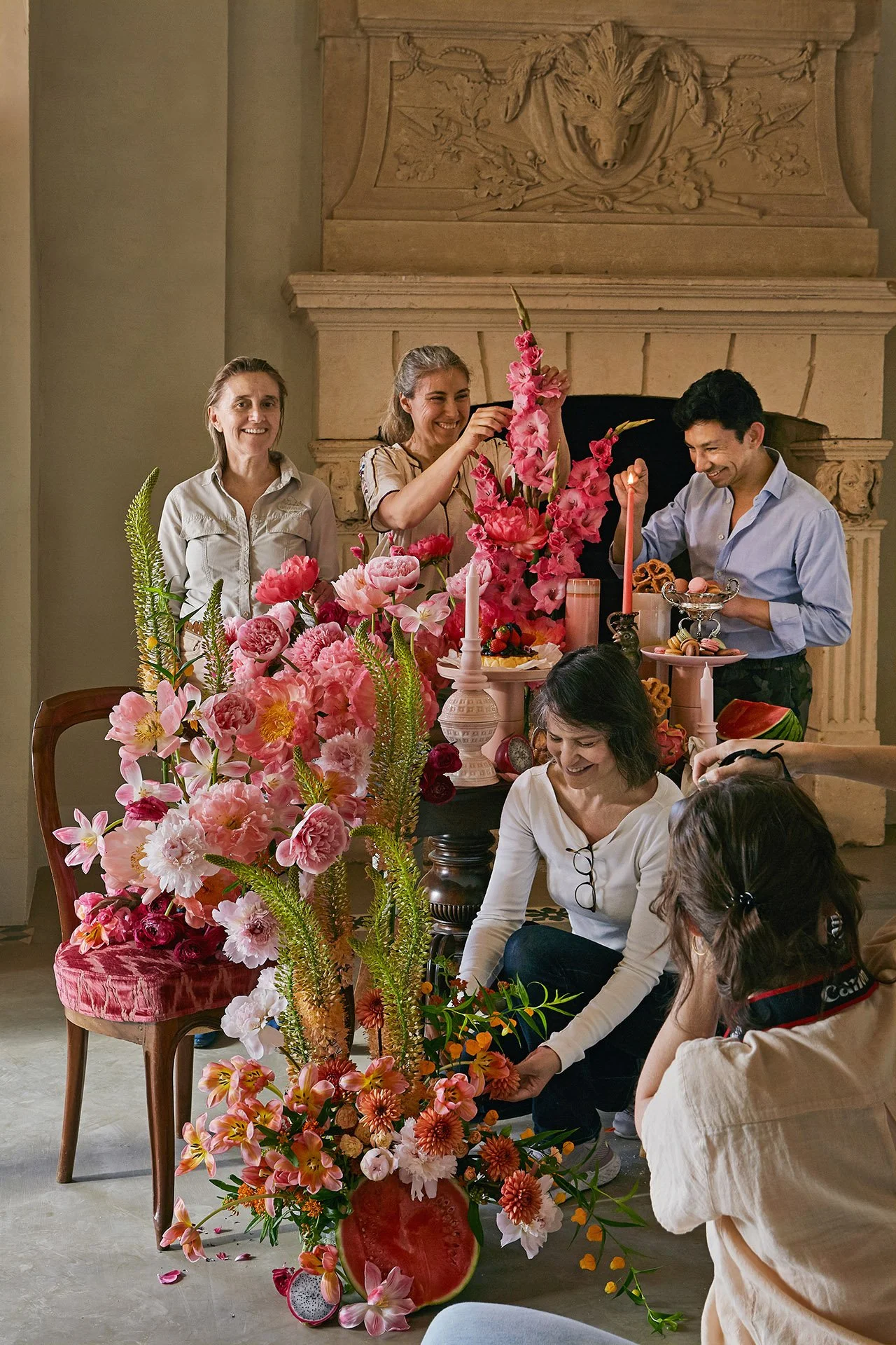 Grupo de personas arreglando flores de colores en una sala elegante con chimenea decorativa al fondo.
