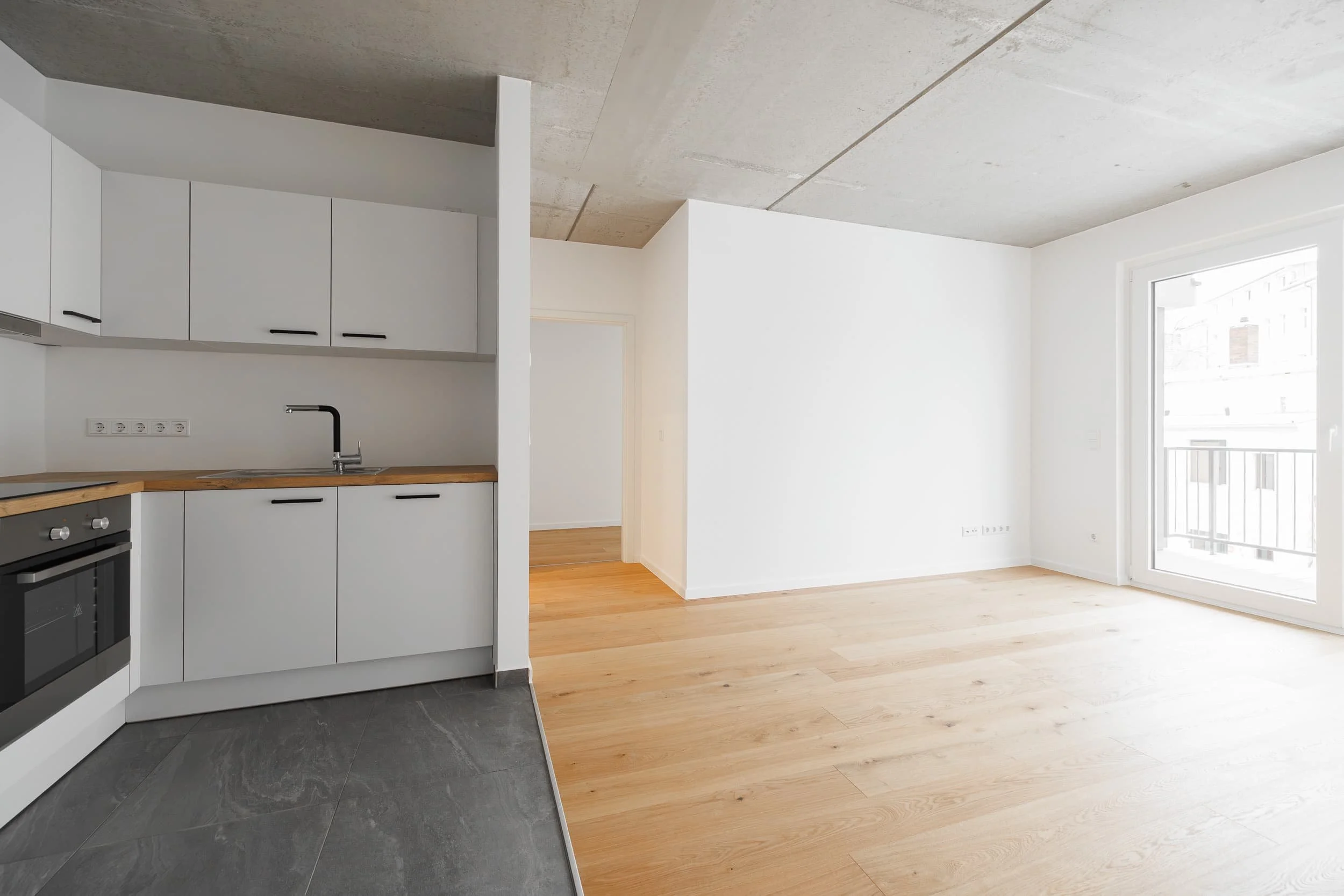 Ein heller, moderner Raum mit einem offenen Grundriss, Holzfußboden und großen Fenstern, die viel Tageslicht hereinlassen. Im Vordergrund eine Küchenzeile mit weißen Schränken und Holzarbeitsplatte, im Hintergrund ein leerer, heller Raum.