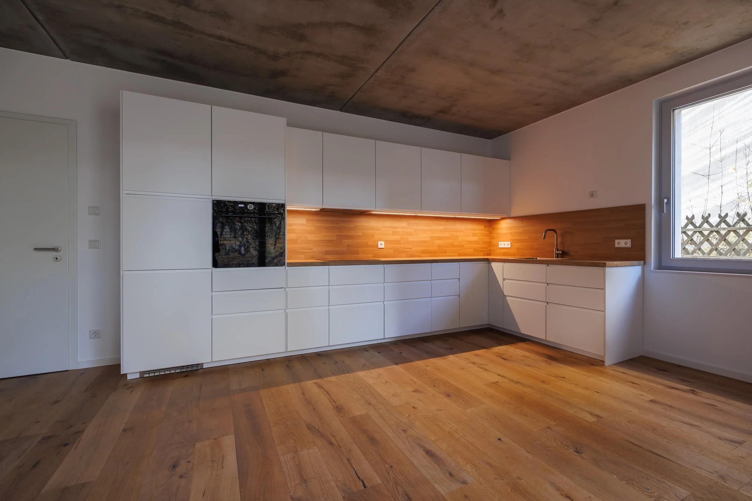 Moderne weiße Einbauküche mit Holzarbeitsplatte, eingebautem Ofen, Spüle und Oberlichtfenster, Holzboden und dunkler Decke.