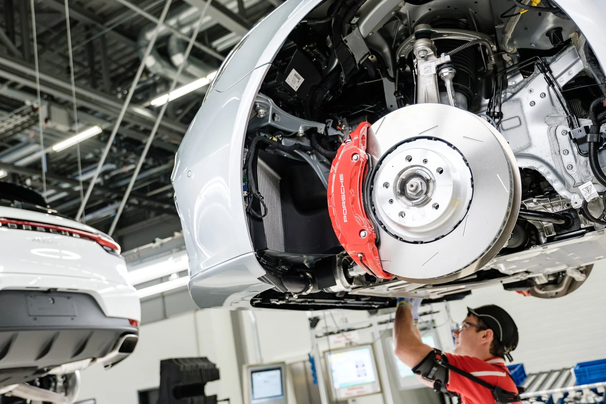 Mitarbeiter arbeitet an einem weißen Auto, das auf einer Wartungsbühne angehoben ist, in einer Autowerkstatt. Das Auto ist teilweise zerlegt, die Bremsanlage ist sichtbar.