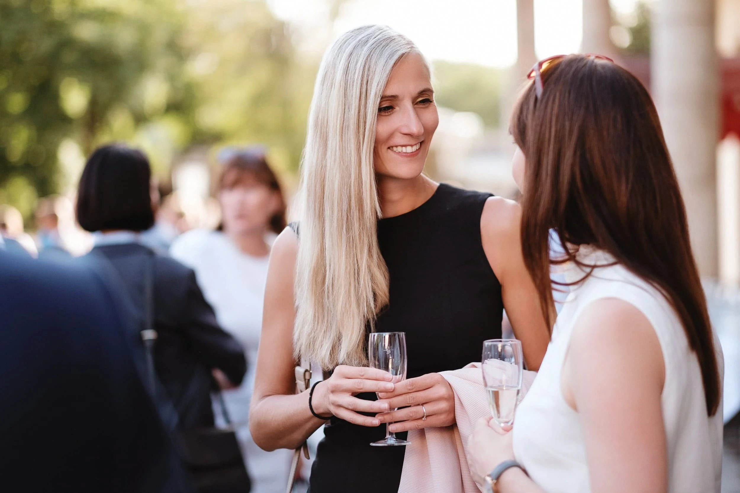 Zwei Frauen unterhalten sich bei einer Veranstaltung im Freien, eine hält ein Glas Sekt in der Hand.