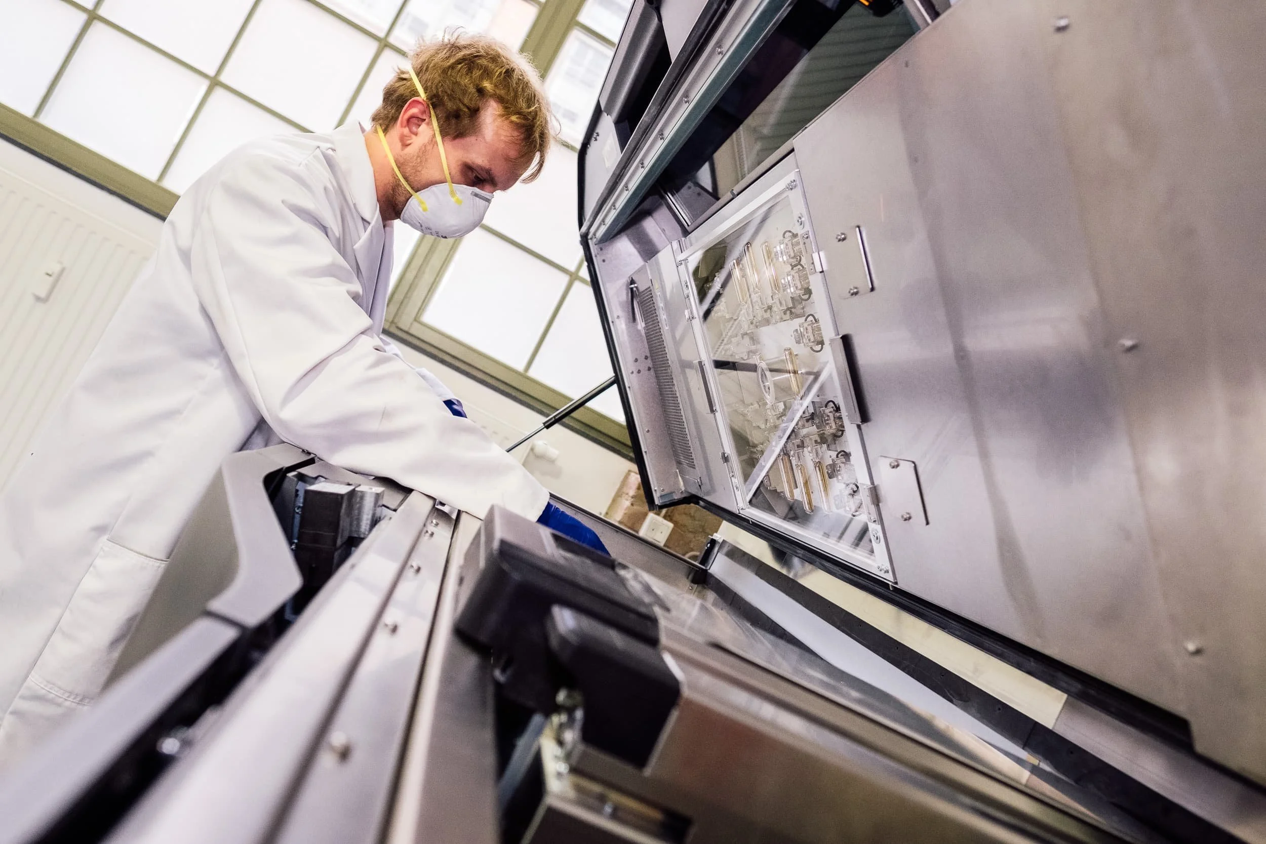Ein Wissenschaftler trägt eine Schutzmaske und arbeitet mit Laborausrüstung in einem Reinraum, umgeben von wissenschaftlichen Instrumenten.