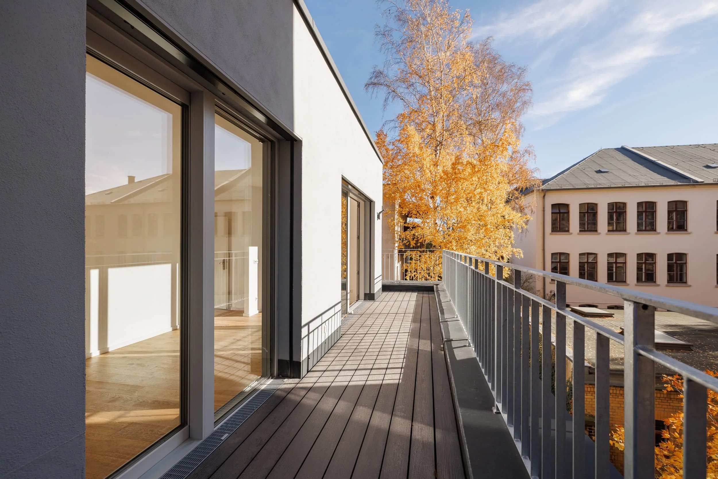 Balkon mit Holzboden und weißen Außenwand, Blick auf einen Baum mit gelben Herbstblättern und eine ältere weiße Gebäudefassade mit großen Fenstern bei sonnigem Himmel.
