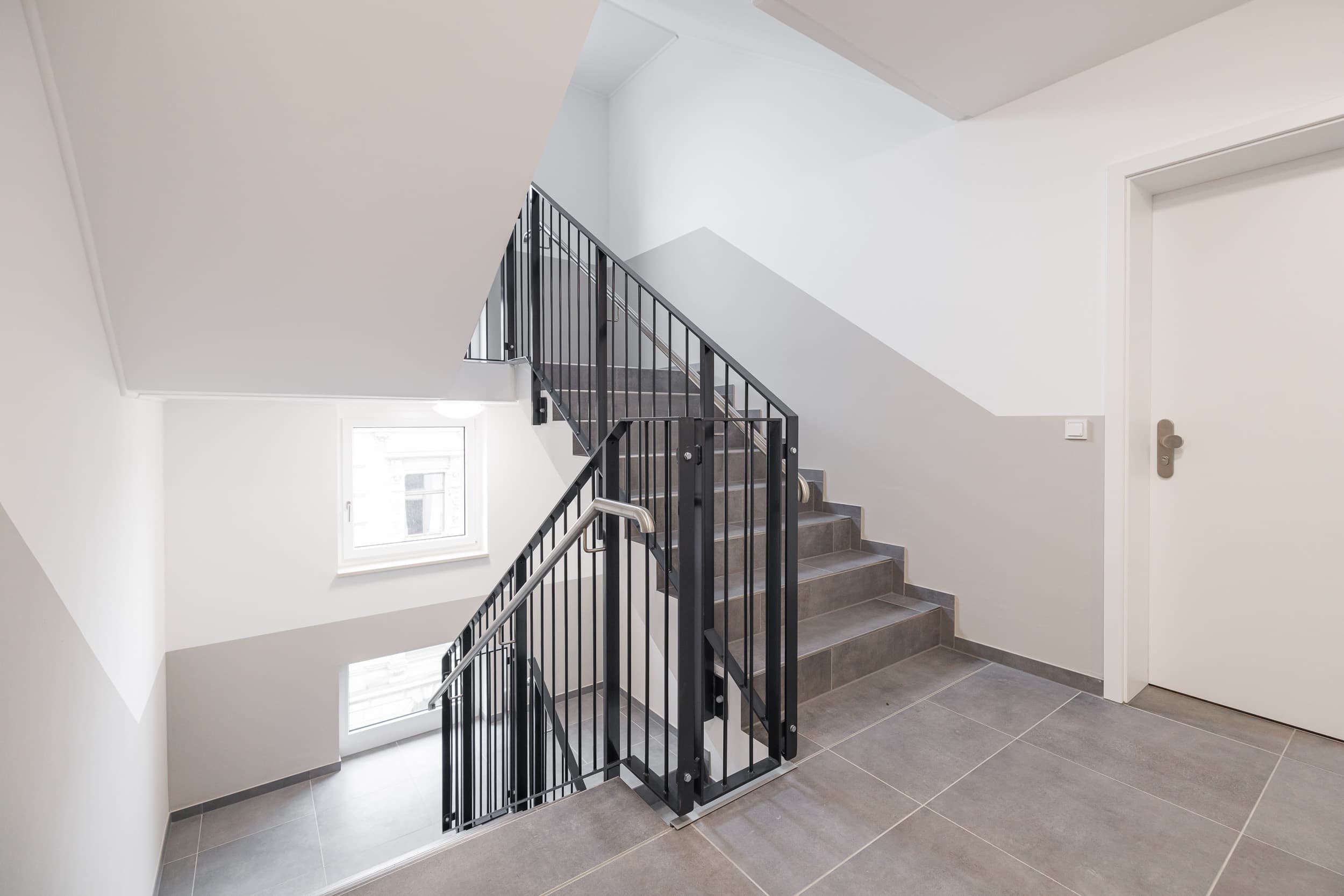 Innenaufnahme eines modernen Treppenhauses mit schwarzen Metallgeländer und grauen Fliesen. Weiß gestrichene Wände und Fenster auf der linken Seite lassen viel Tageslicht herein.