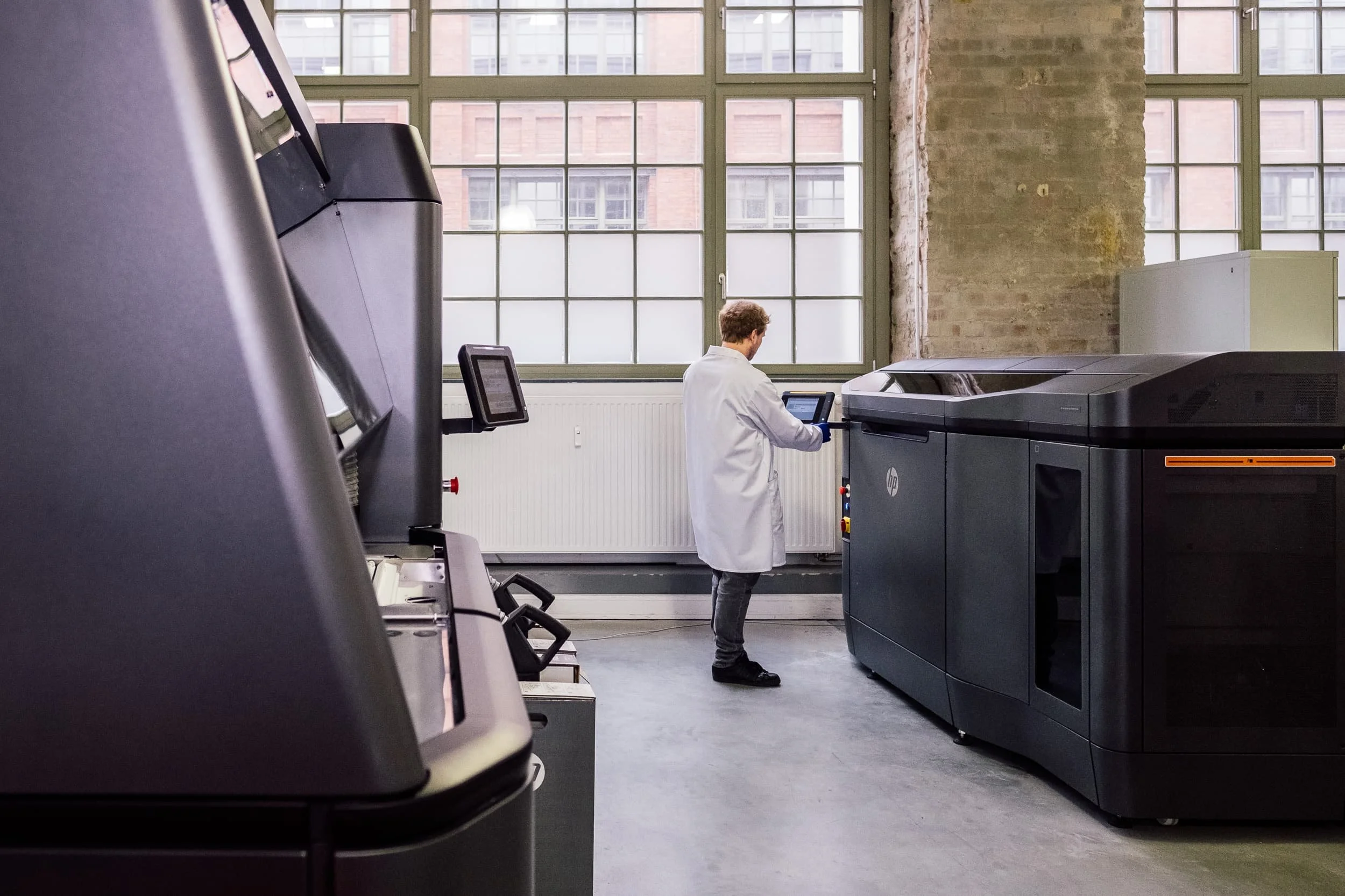 Ein Mann in einem weißen Kittel steht vor einem großen Drucker in einem modernen, industrielichen Raum mit großen Fenstern und einer rauen Wand.