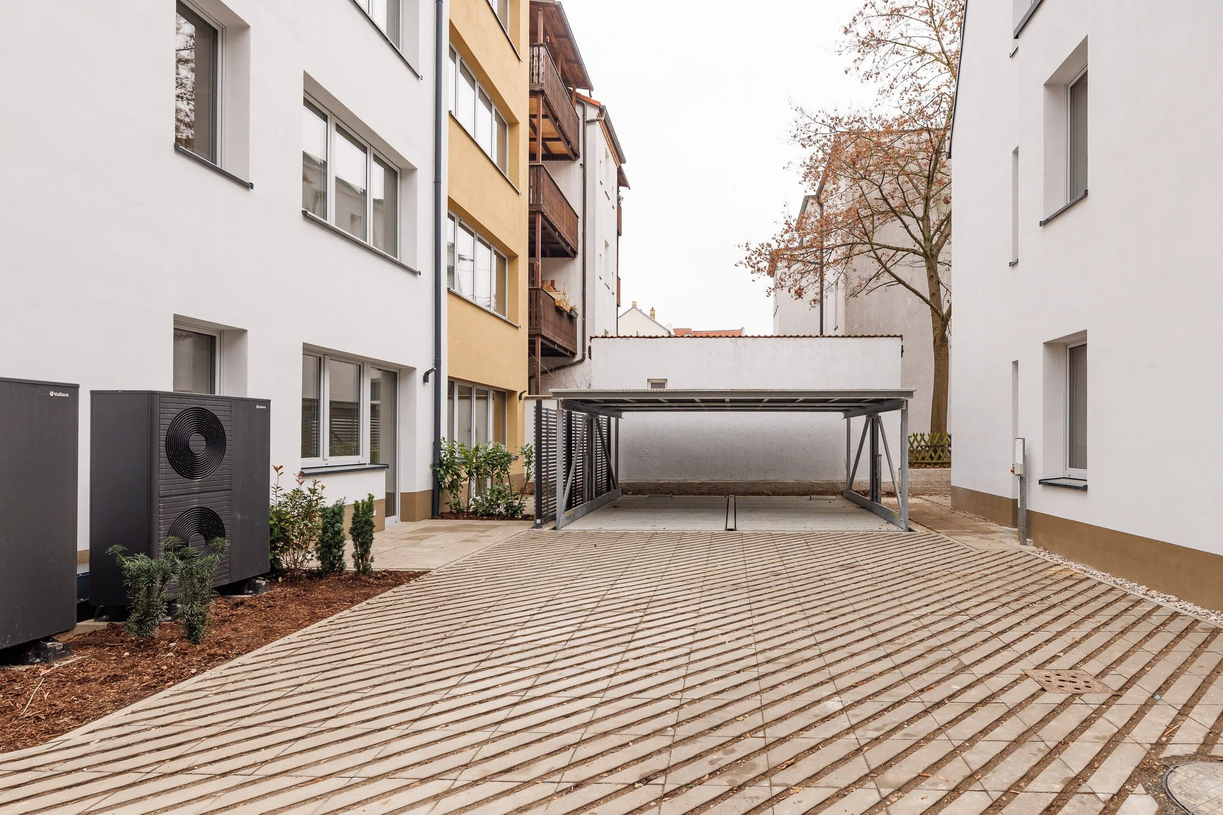 Bahnhofshof mit gepflastertem Boden, moderne Mehrfamilienhäuser mit weißen und gelben Fassaden, Balkone, ein Baum mit Herbstlaub, Heizgeräte im Vordergrund, ein Carport, im Hintergrund ein graues Himmel.