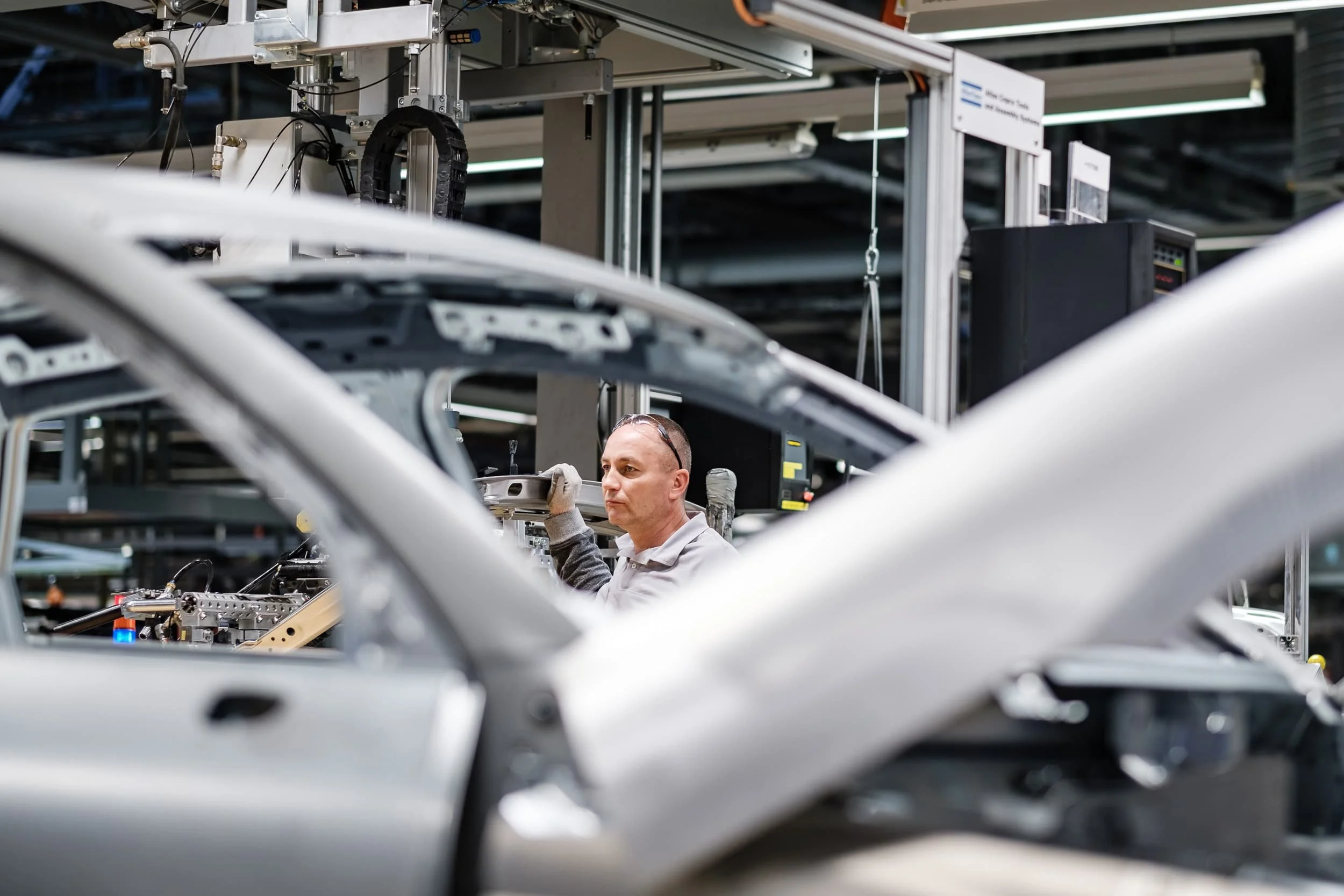 Mann arbeitet an einem technischen Gerät in einer Fertigungshalle, umgeben von Metallrahmen und industriellen Maschinen.
