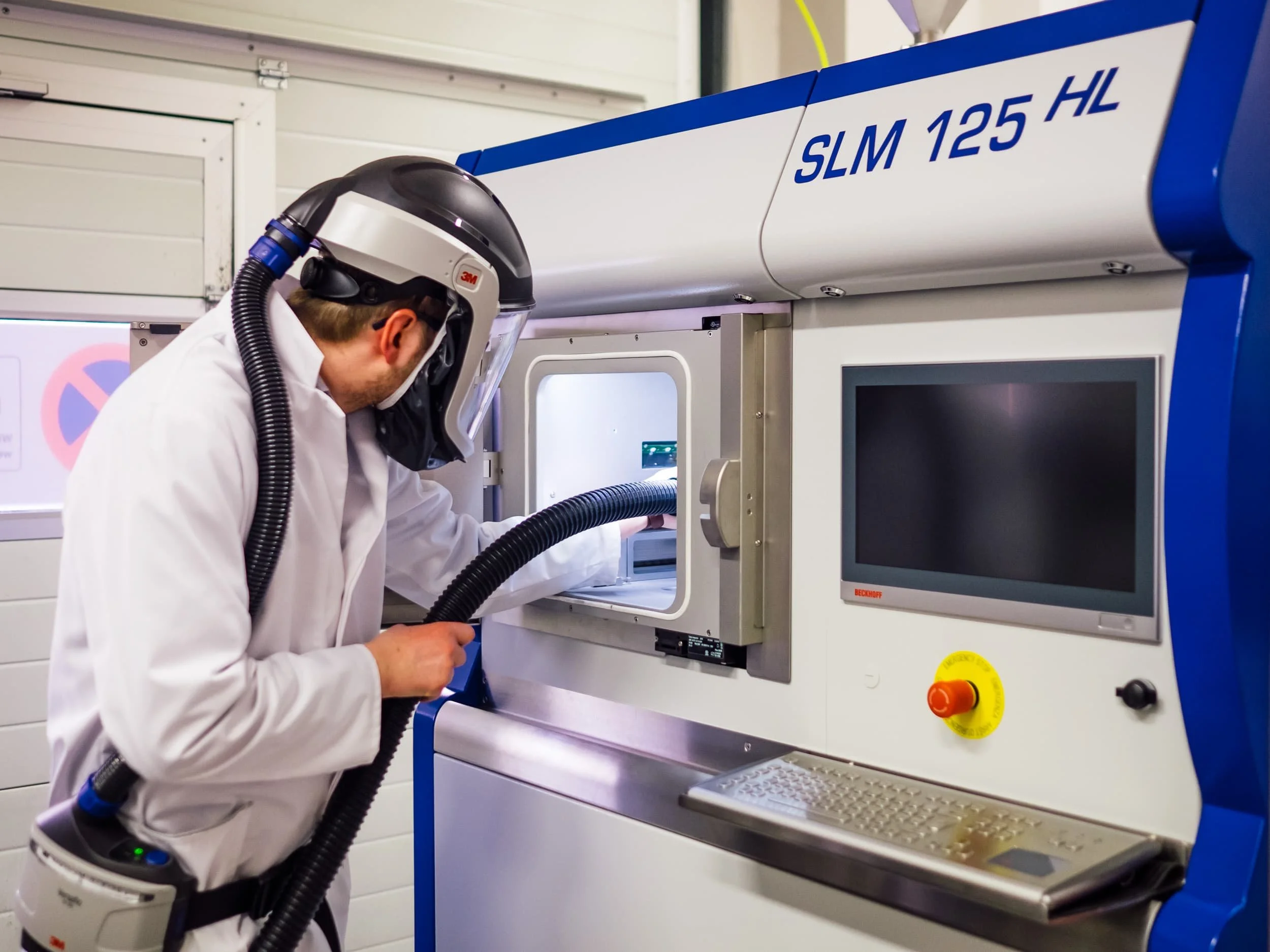 Ein Mensch in Schutzkleidung arbeitet mit einem Atemschutz in einer technischen oder laborbezogenen Umgebung, nahe einem Gerät mit Monitor und Schaltflächen.