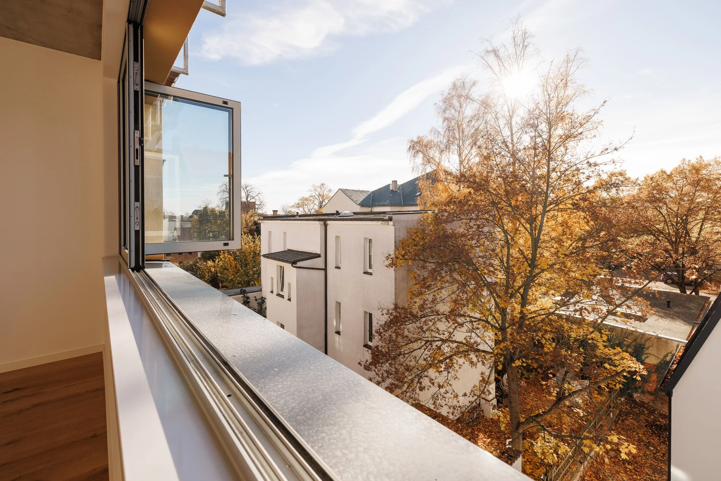 Ein offenes Fenster in einer Wohnung mit Blick auf eine Straße mit Gebäuden und Bäumen im Herbst bei Sonnenlicht.