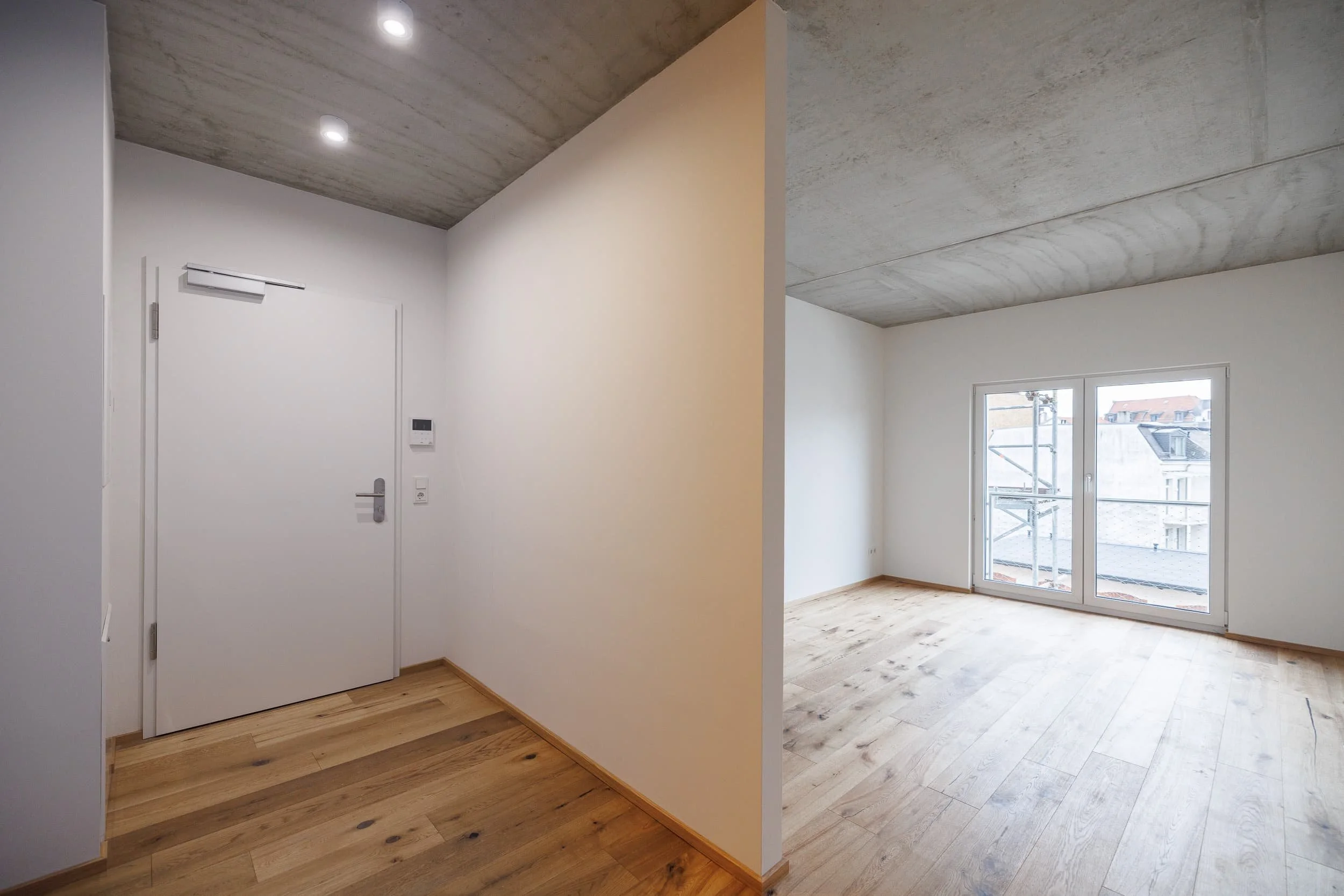 Innenraum eines unmöblierten Zimmers mit Holzfußboden, großen Fenstern und weißen Wänden, mit Sicht auf ein Nachbargebäude durch die Fenster.