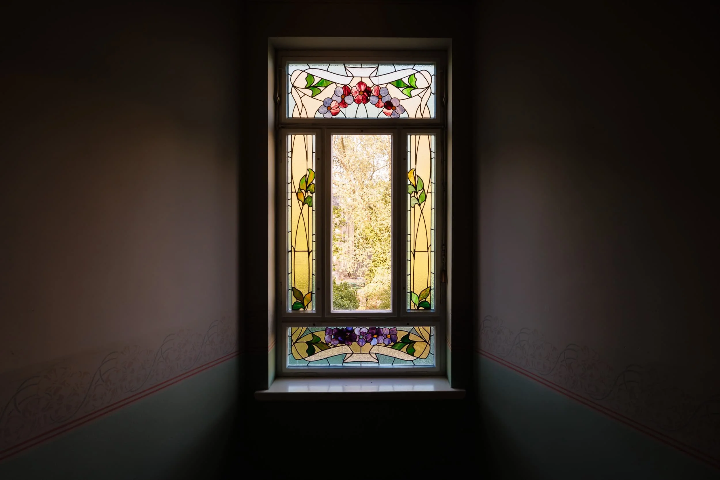 Bunt verziertes Fenster mit Blumenmotiven und einer Aussicht auf Bäume draußen, im dunklen Raum.