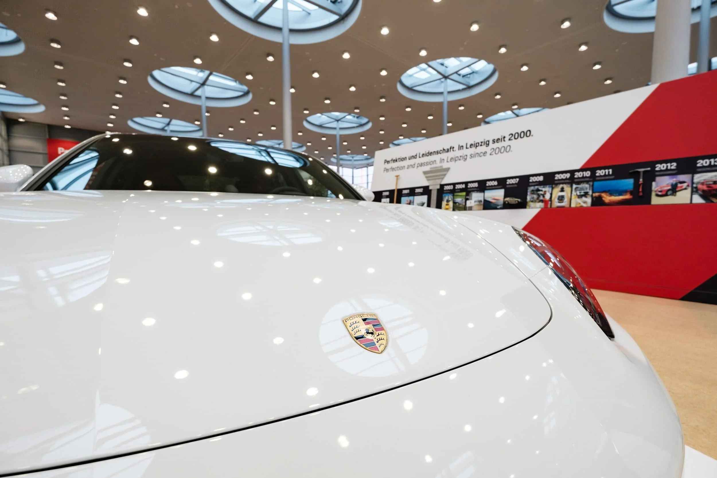 Ein weißes Porsche-Auto, das im Ausstellungsraum präsentiert wird, mit dem Porsche-Logo auf der Motorhaube, im Hintergrund eine Informationswand mit Timeline.
