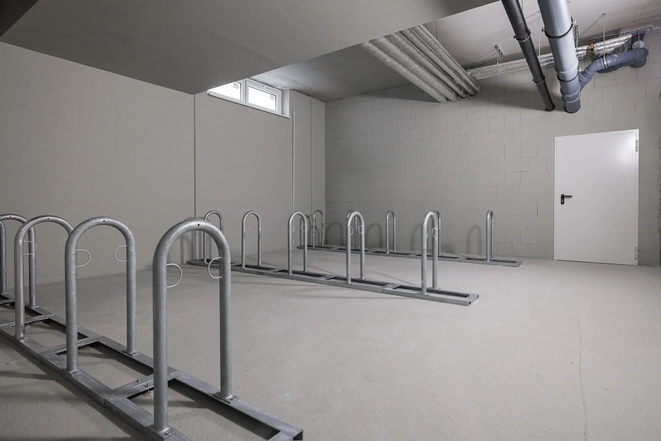 Ein leerer Raum in einem Gebäude mit weißen Wänden, Decke und Tür, sowie Fahrradständern im Vordergrund und Rohrleitungen an der Decke.