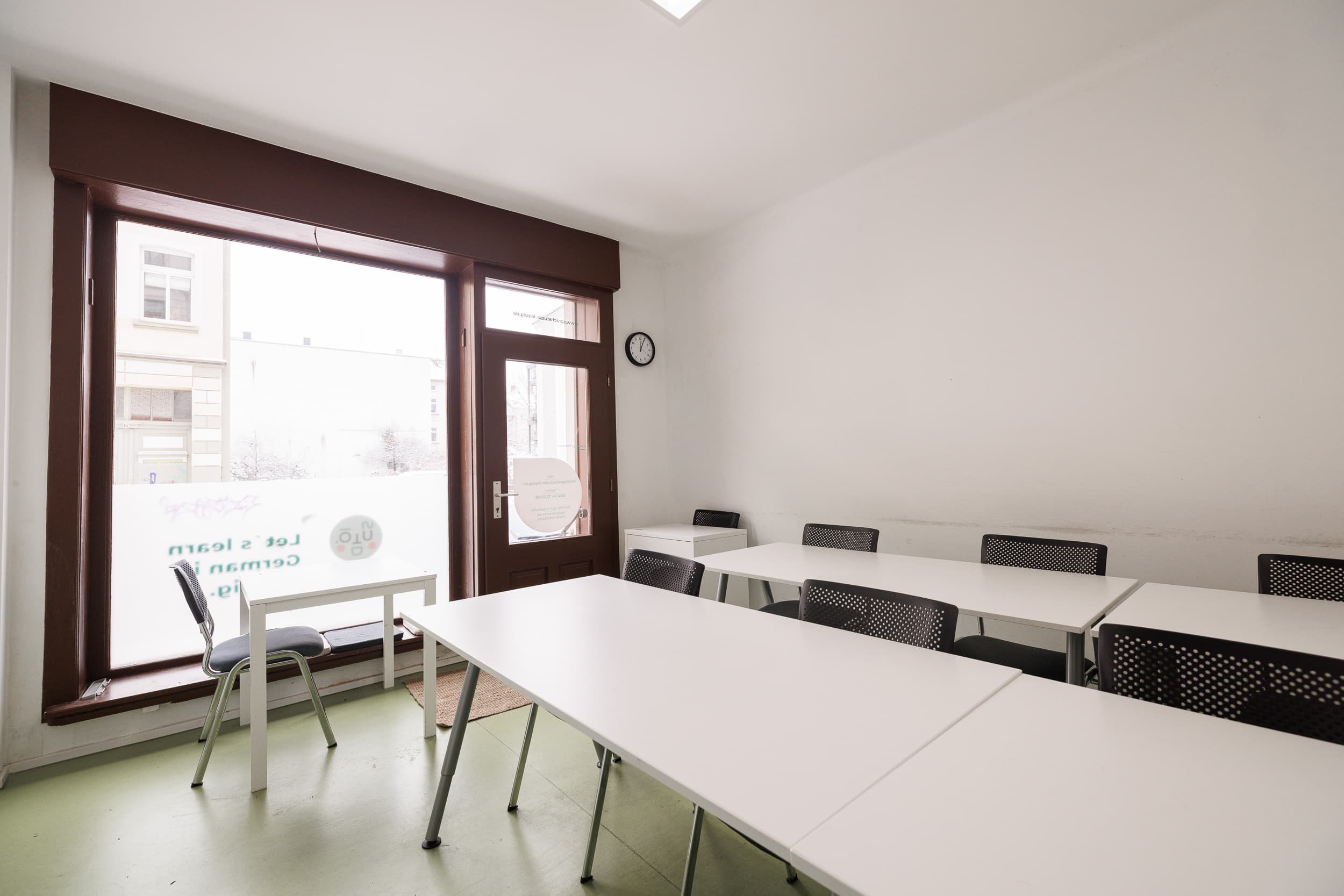 Ein heller Raum mit weißen Wänden, mehreren weißen Tischen und schwarzen Stühlen. Große Fenster mit dunklen Rahmen lassen natürliches Licht herein, und es ist ein Wanduhr auf der Wand neben den Fenstern sichtbar.