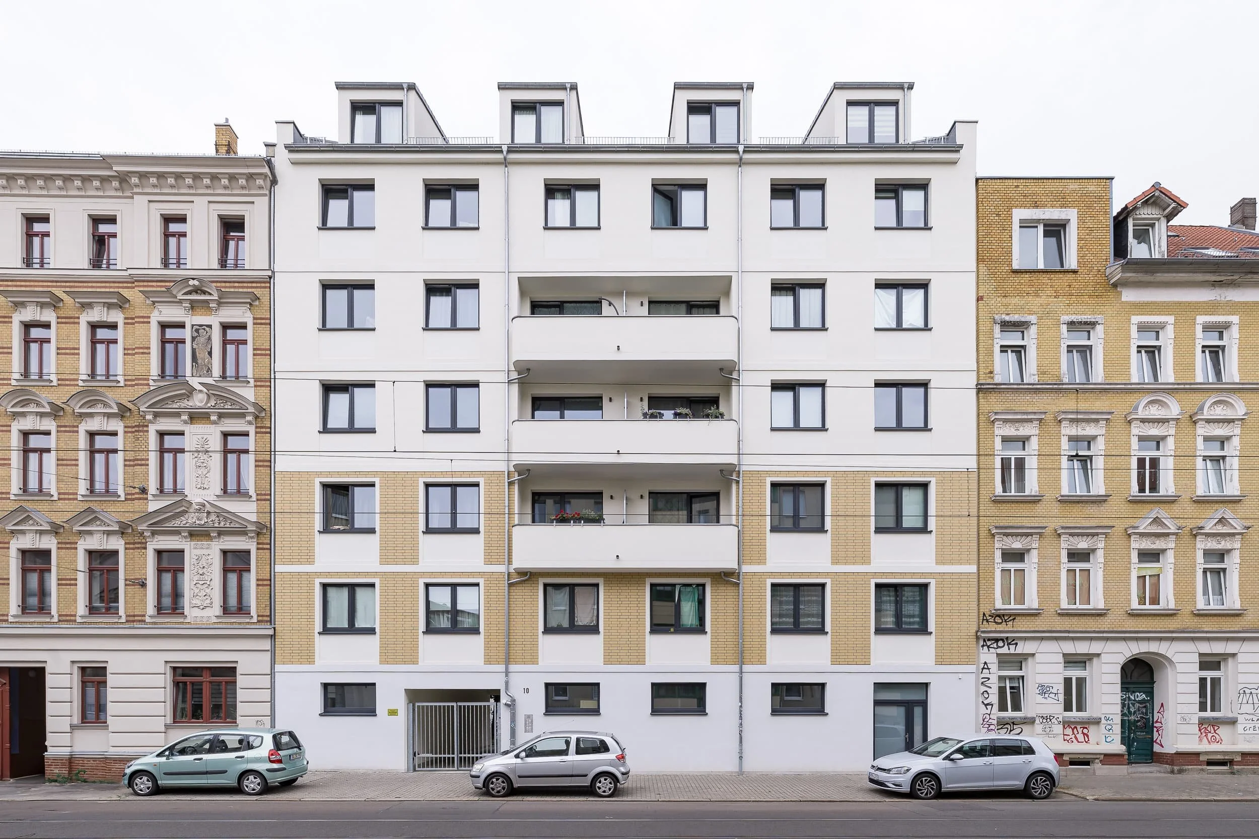 Mehrstöckige Wohnhäuser in einer Stadt, darunter ein modernes weißes Gebäude zwischen zwei historisch aussehenden Gebäuden, mit geparkten Autos an der Straße davor.