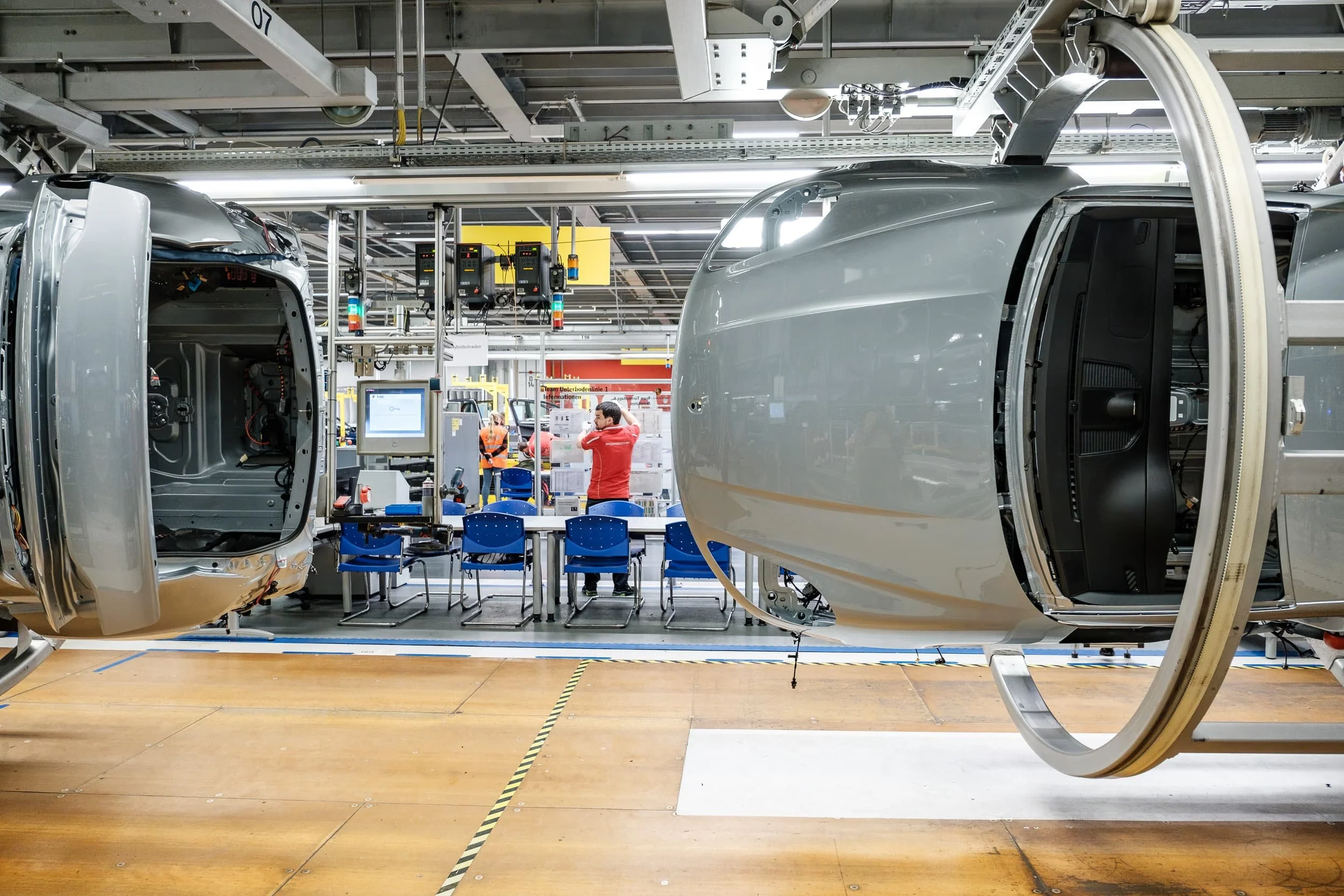Innenansicht eines Automobilwerks mit zwei Fahrzeugkarosserien im Bau, mehrere Arbeiter und technische Geräte im Hintergrund.