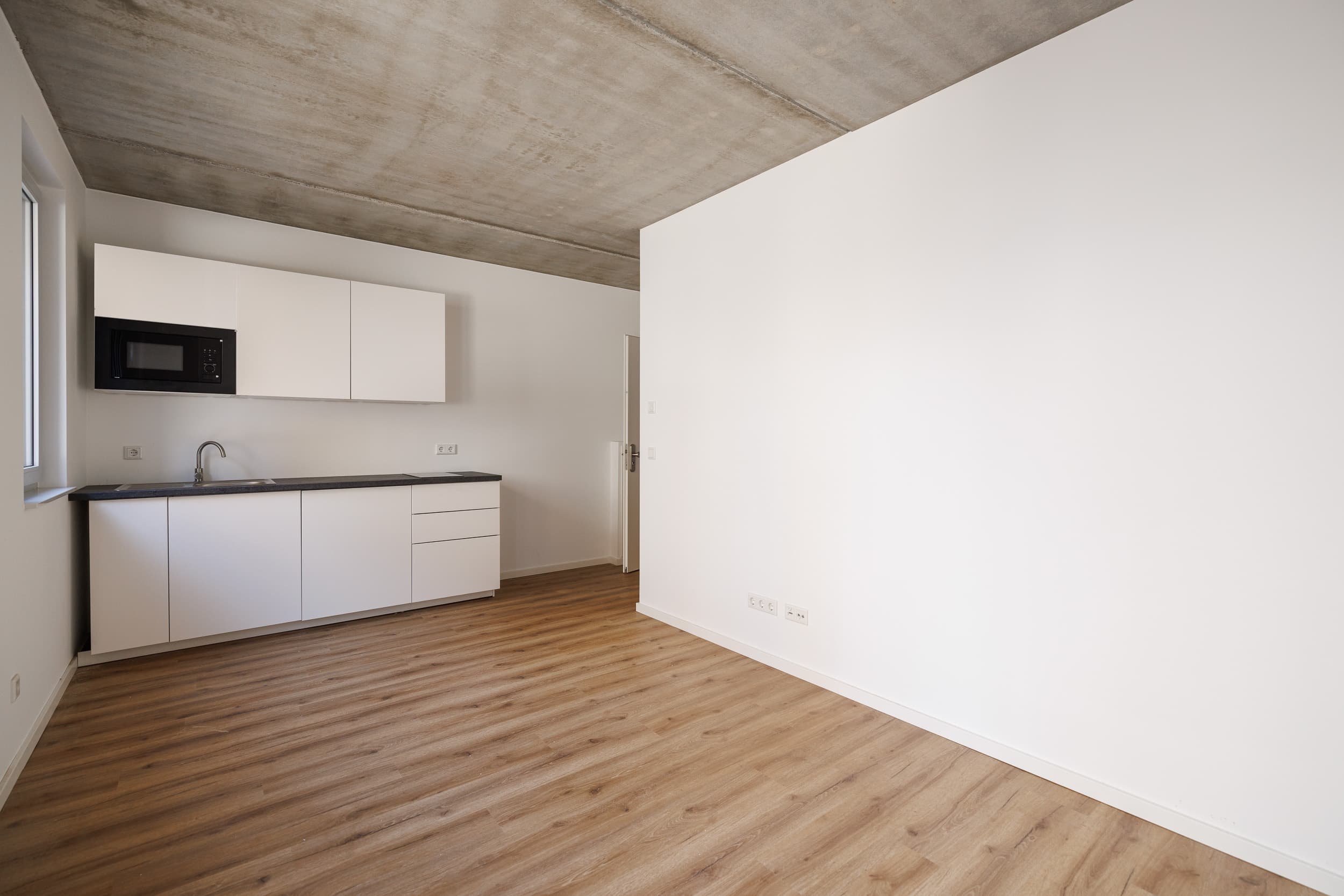 Leerer Küchenraum mit weißen Schränken, schwarzen Arbeitsplatten, Spüle, Mikrowelle, Holzboden und schräg verlaufender Decke.