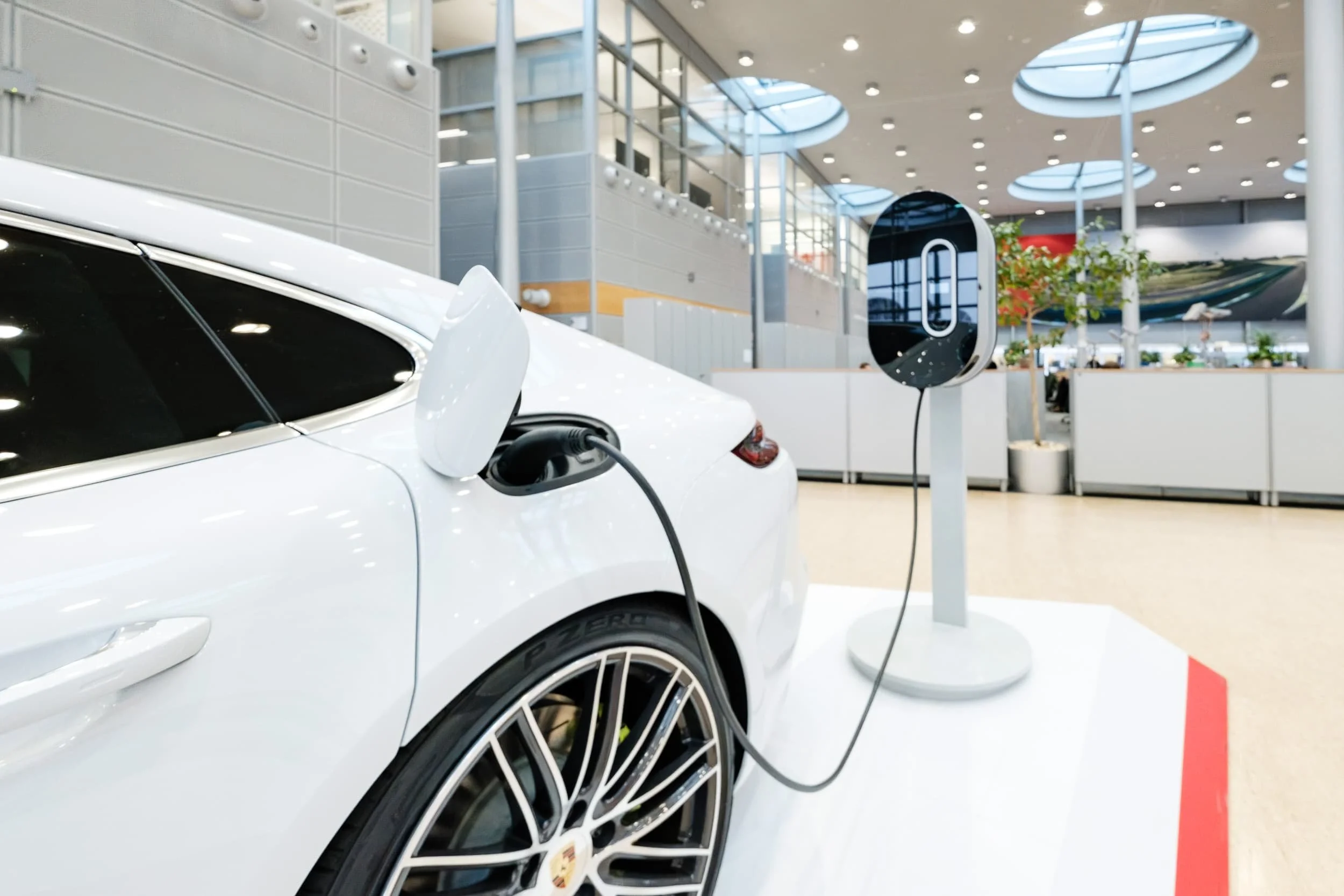 Ein weißes Elektroauto wird aufgeladen, mit einem Ladekabel verbunden, in einem modernen Gebäude mit viel natürlichem Licht, Pflanzen und großen Fenstern.