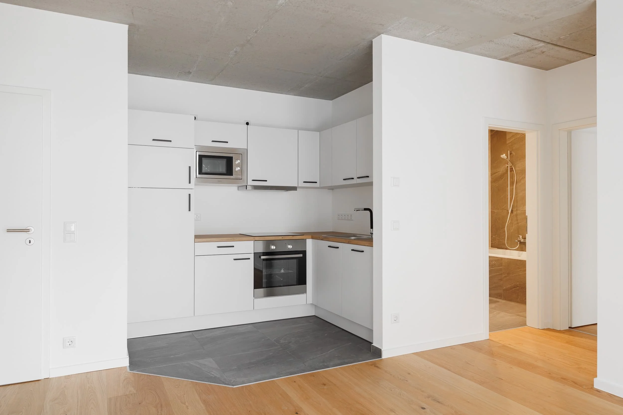 Moderne weiße Küchenschränke mit dunklen Griffen, eingebauter Mikrowelle, Herd und Ofen, offener Grundriss mit Holz- und Fliesenboden, Badezimmer mit Dusche im Hintergrund.