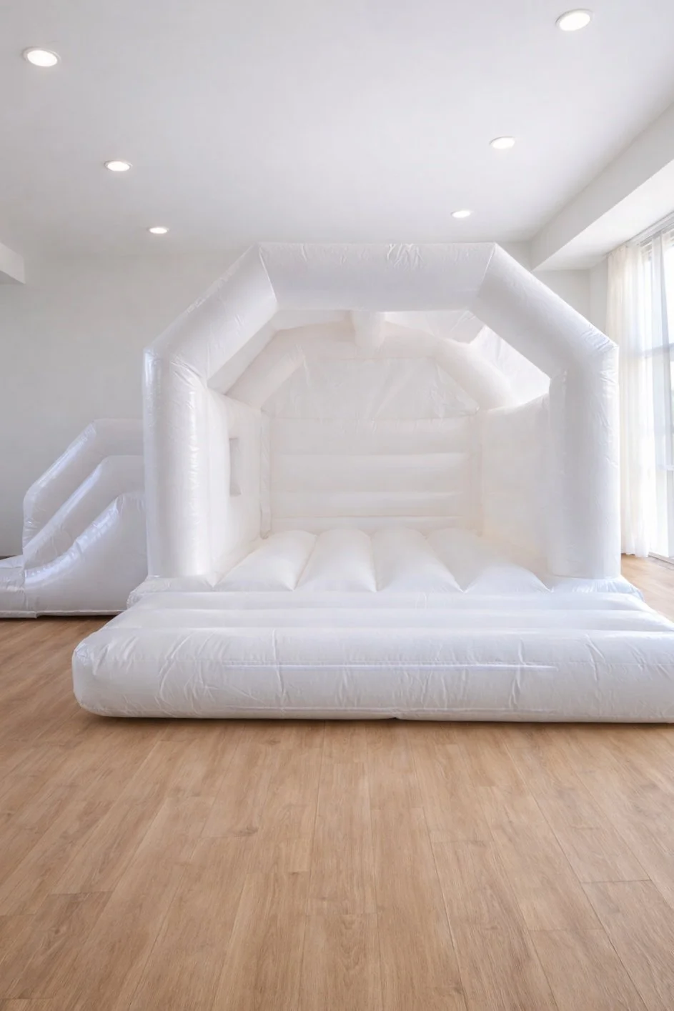 Indoor inflatable white house with an inflatable slide on the side, set up on a wooden floor with a bright room and large window with curtains in the background.