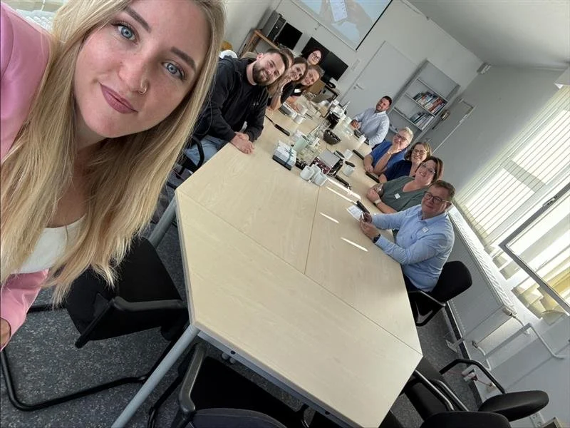 Sieben Menschen sitzen um einen Konferenztisch in einem Büro, einige schauen in die Kamera, der Raum ist gut erleuchtet mit großen Fenstern.