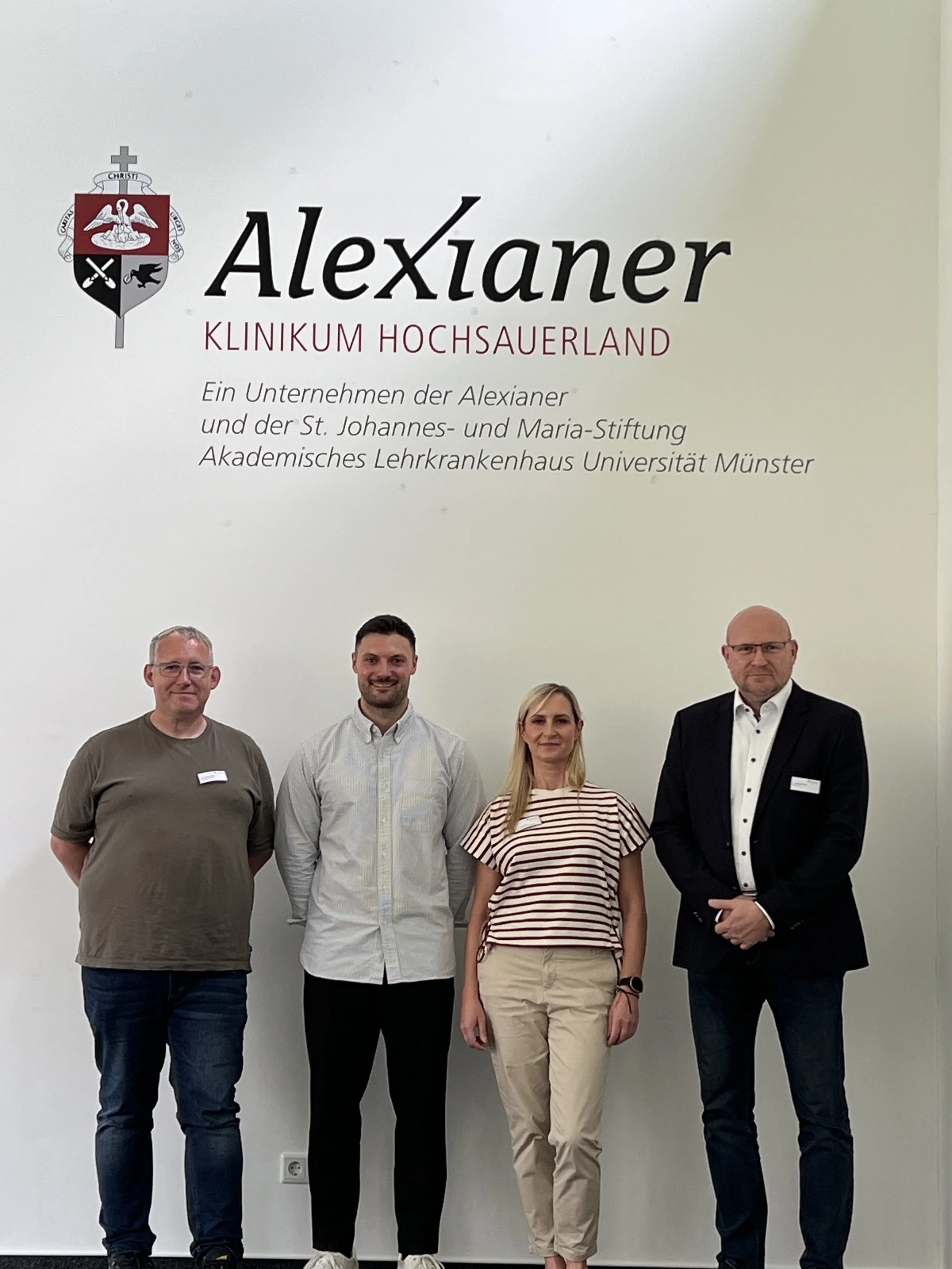 Vier Personen stehen vor einer weißen Wand mit einem Schriftzug und einem Wappen, darunter steht: 'Alexianer Klinikum Hochsauerland'.