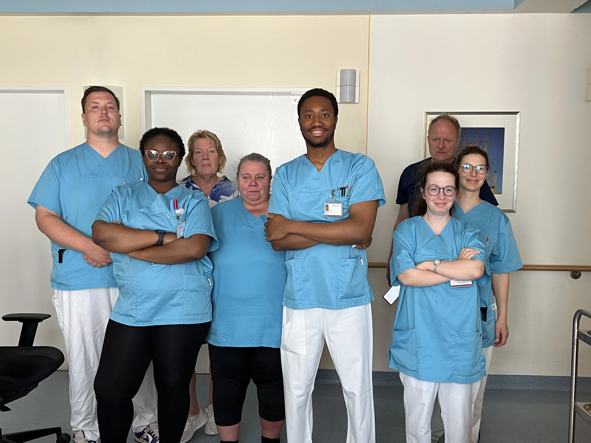Eine Gruppe von acht medizinischen Fachkräften, bestehend aus Ärzten und Krankenschwestern, posiert in einem Krankenhausflur. Sie tragen blaue OP-Kleidung, einige haben Namensschilder und medizinisches Zubehör an ihrem Outfit.
