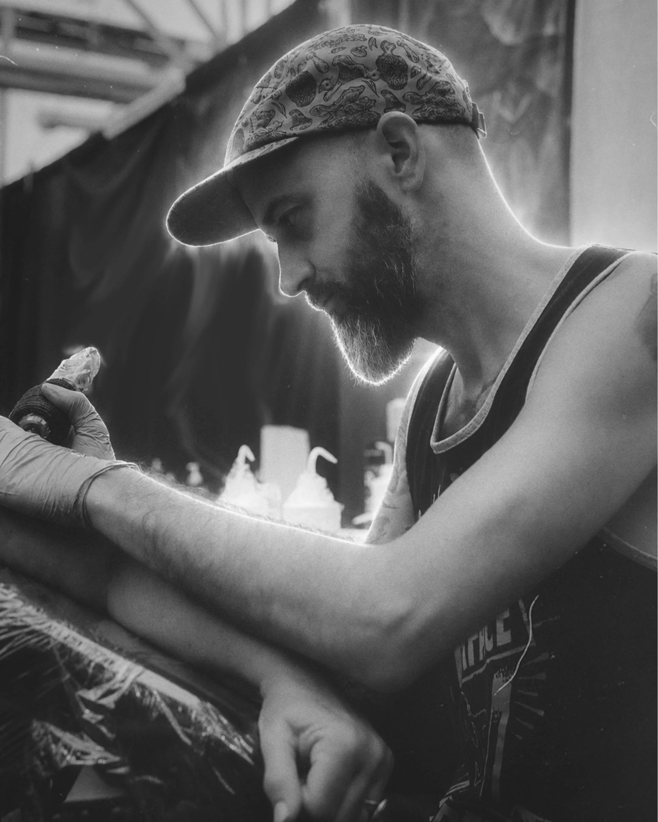 A man with a checkered baseball cap, a beard, and a sleeveless shirt is tattooing someone. The scene is in black and white, and it appears to be in a tattoo studio.