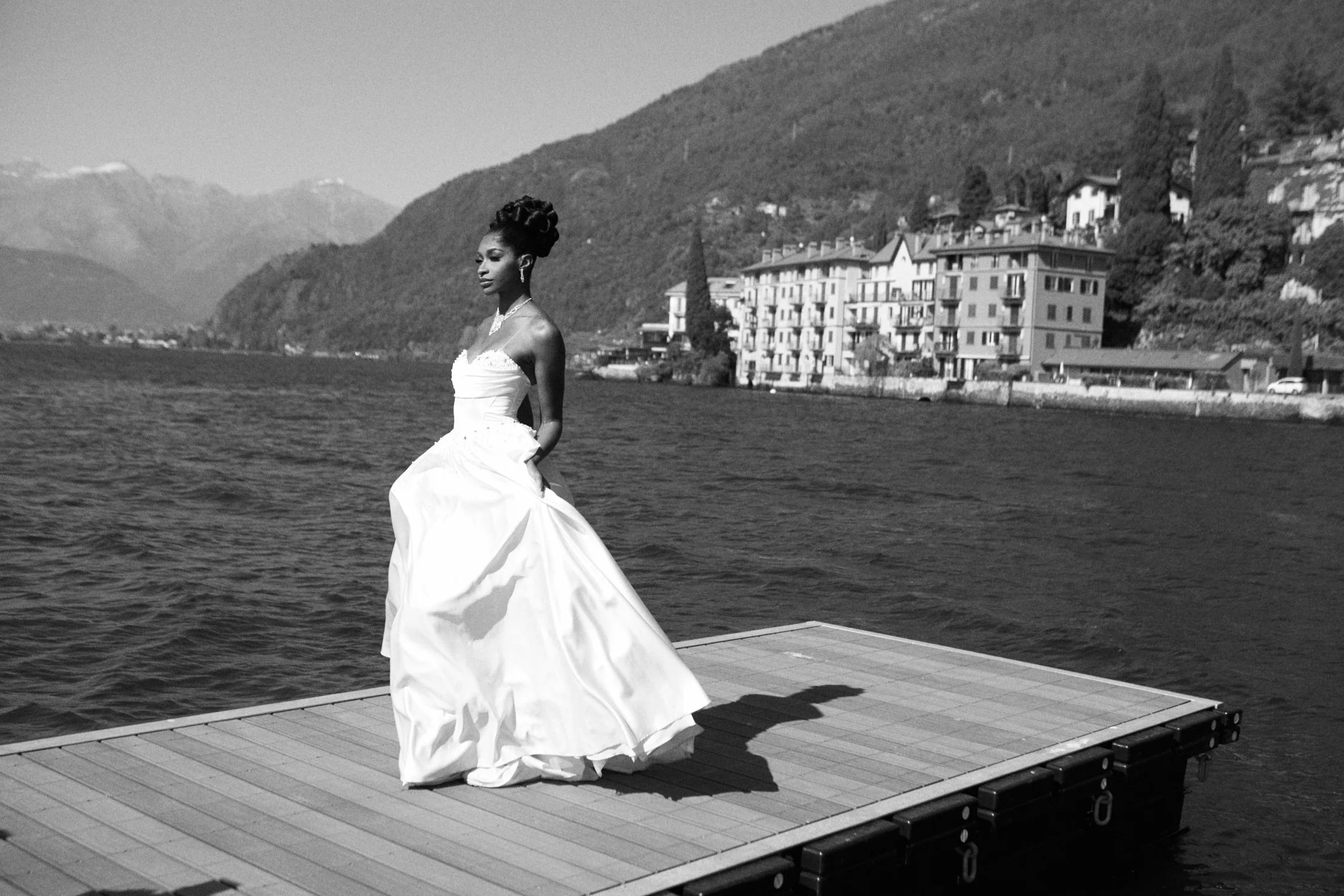 A woman in a strapless wedding dress with a full skirt stands on a wooden dock by the water, with hills and buildings in the background.