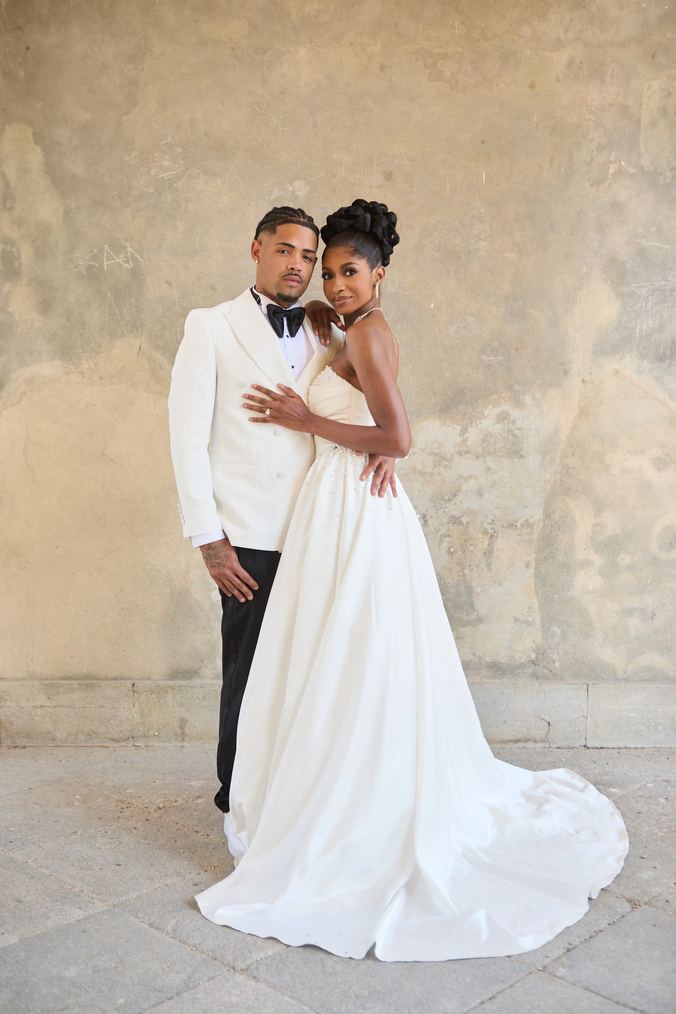 A man and woman in formal attire standing close against a worn concrete wall. The man is wearing a white tuxedo jacket with black bow tie and black pants, with his hand on the woman's waist. The woman is in a strapless white wedding gown with her hai