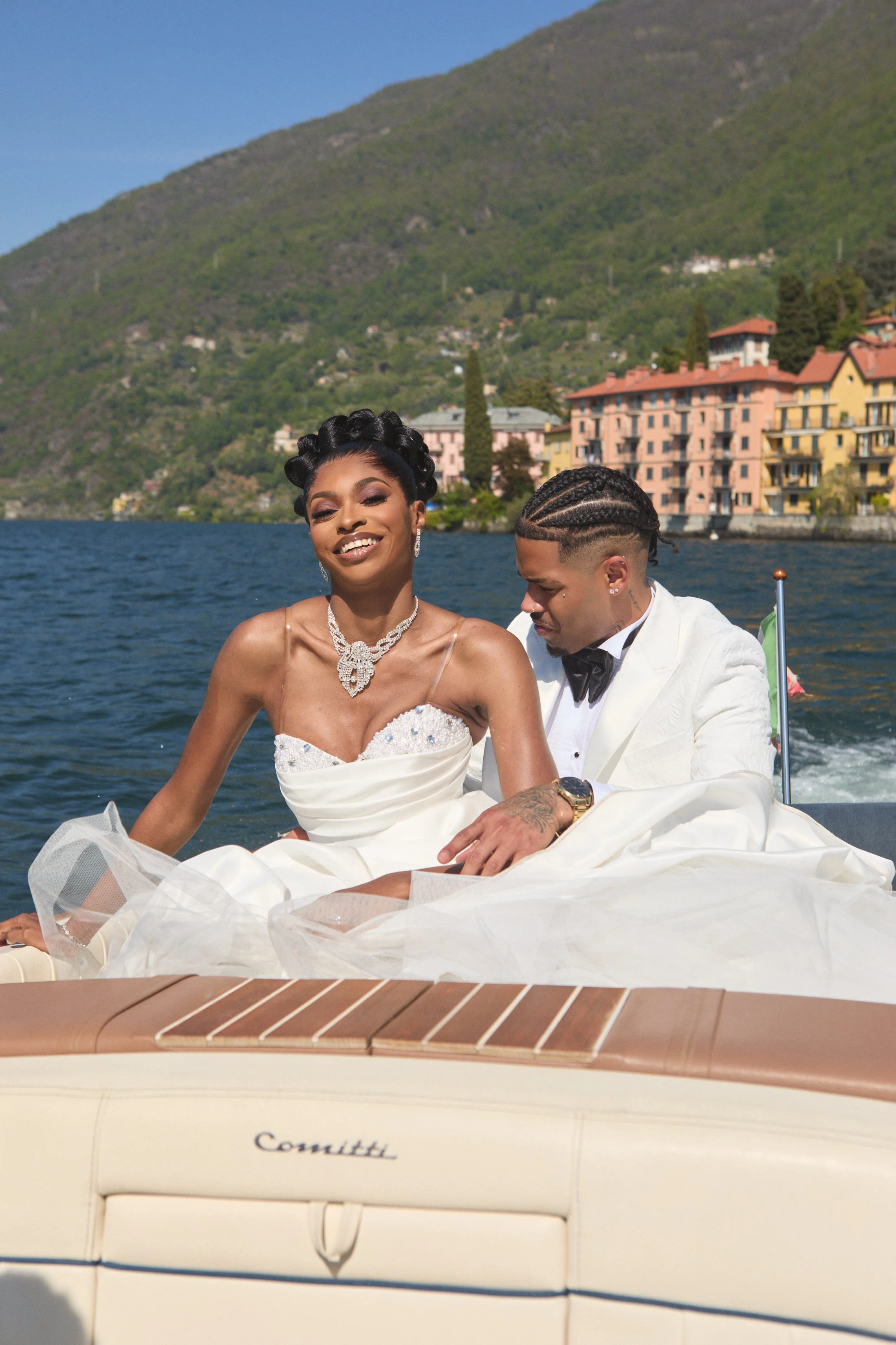 A bride and groom celebrating on a boat on a lake with hills and colorful buildings in the background. The bride is smiling and wearing a white wedding dress with jewelry, and the groom is looking down, dressed in a white tuxedo.