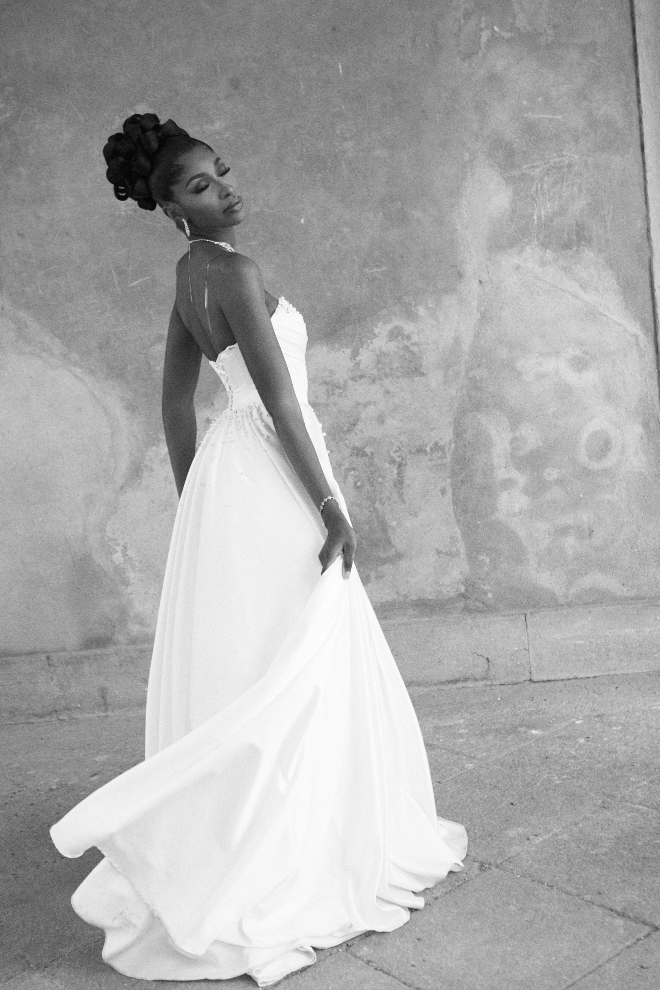 A woman in a white wedding dress with a strapless top and flowing skirt, standing against a textured gray wall. Her eyes are closed and she has an intricate updo hairstyle, accessorized with earrings and a necklace.