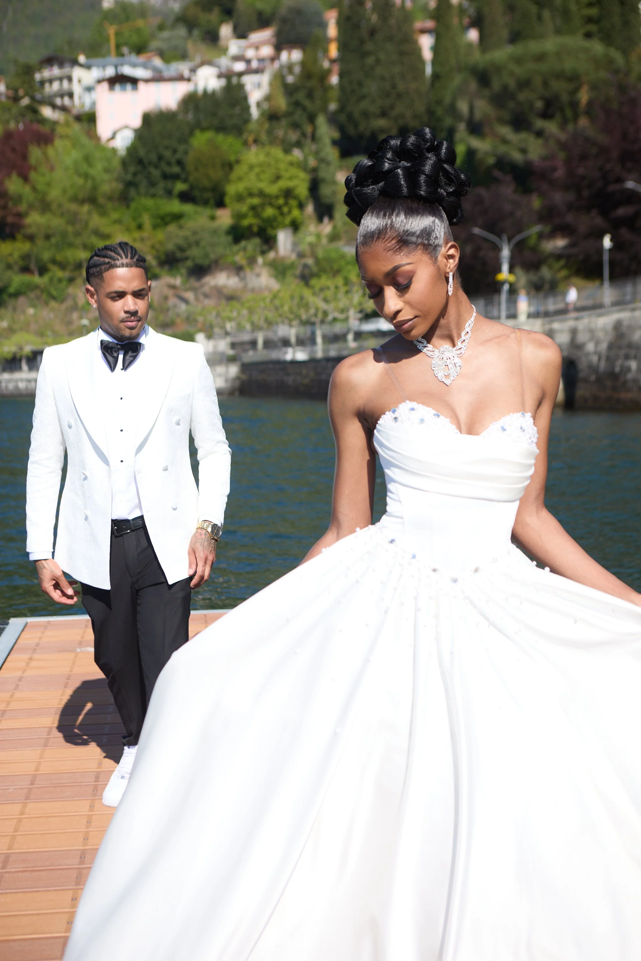 A woman in a white wedding gown with an intricate black hairdo and jewelry, standing on a dock by a body of water, with a man in a white tuxedo and black bow tie standing behind her. There are colorful autumn trees and houses in the background.