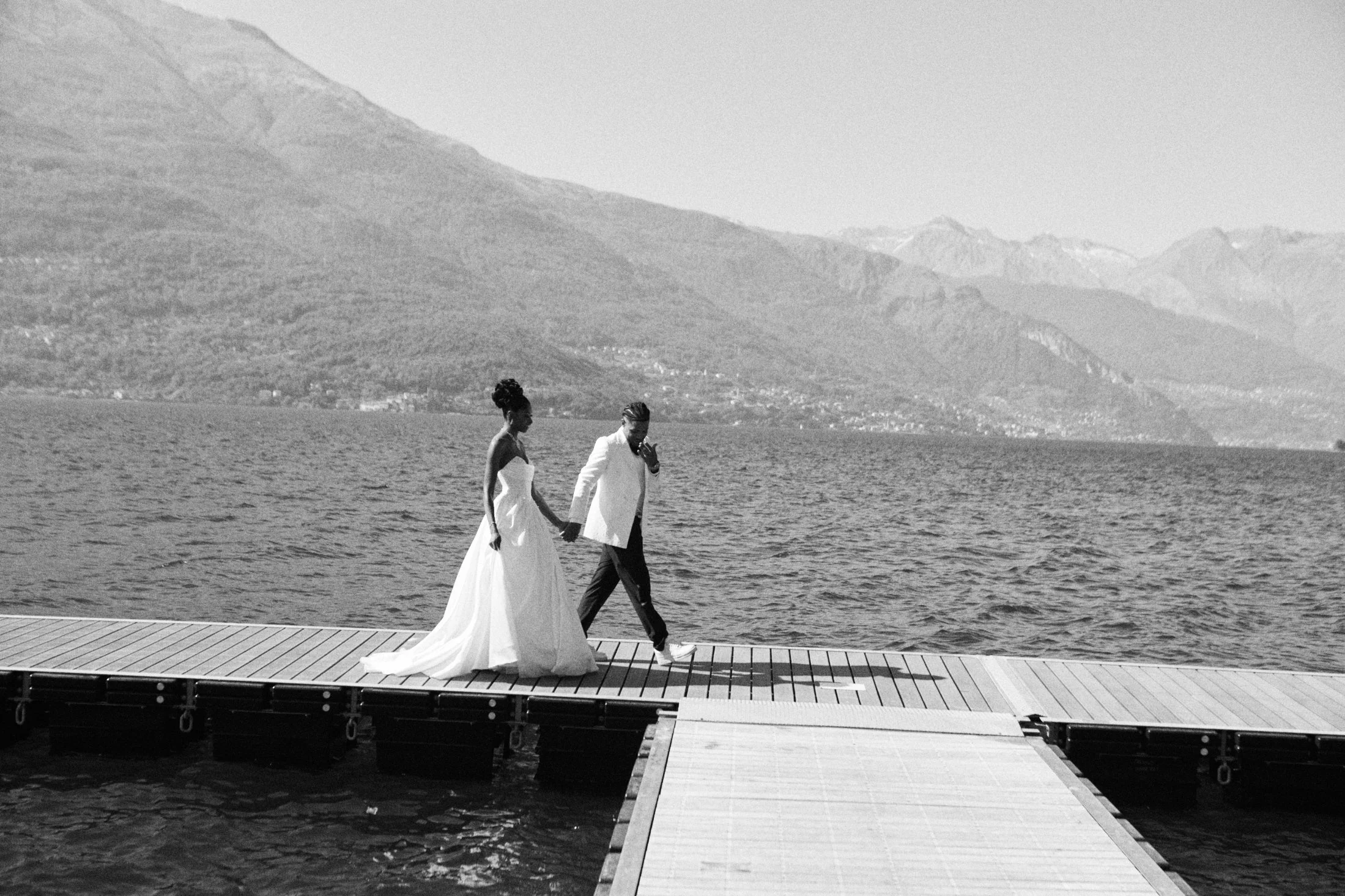 A couple in wedding attire walking hand in hand on a dock by a large body of water with mountains in the background.